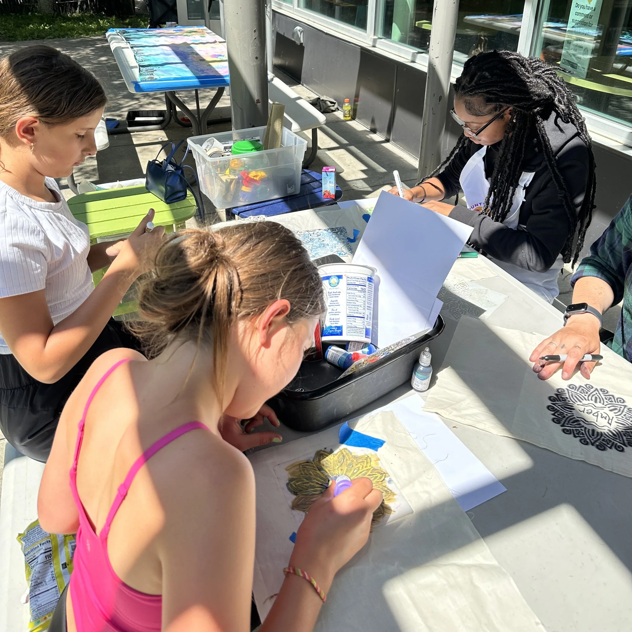 Children and adults participating in an outdoor art activity, drawing and coloring at a table with supplies, under a partly sunny sky.