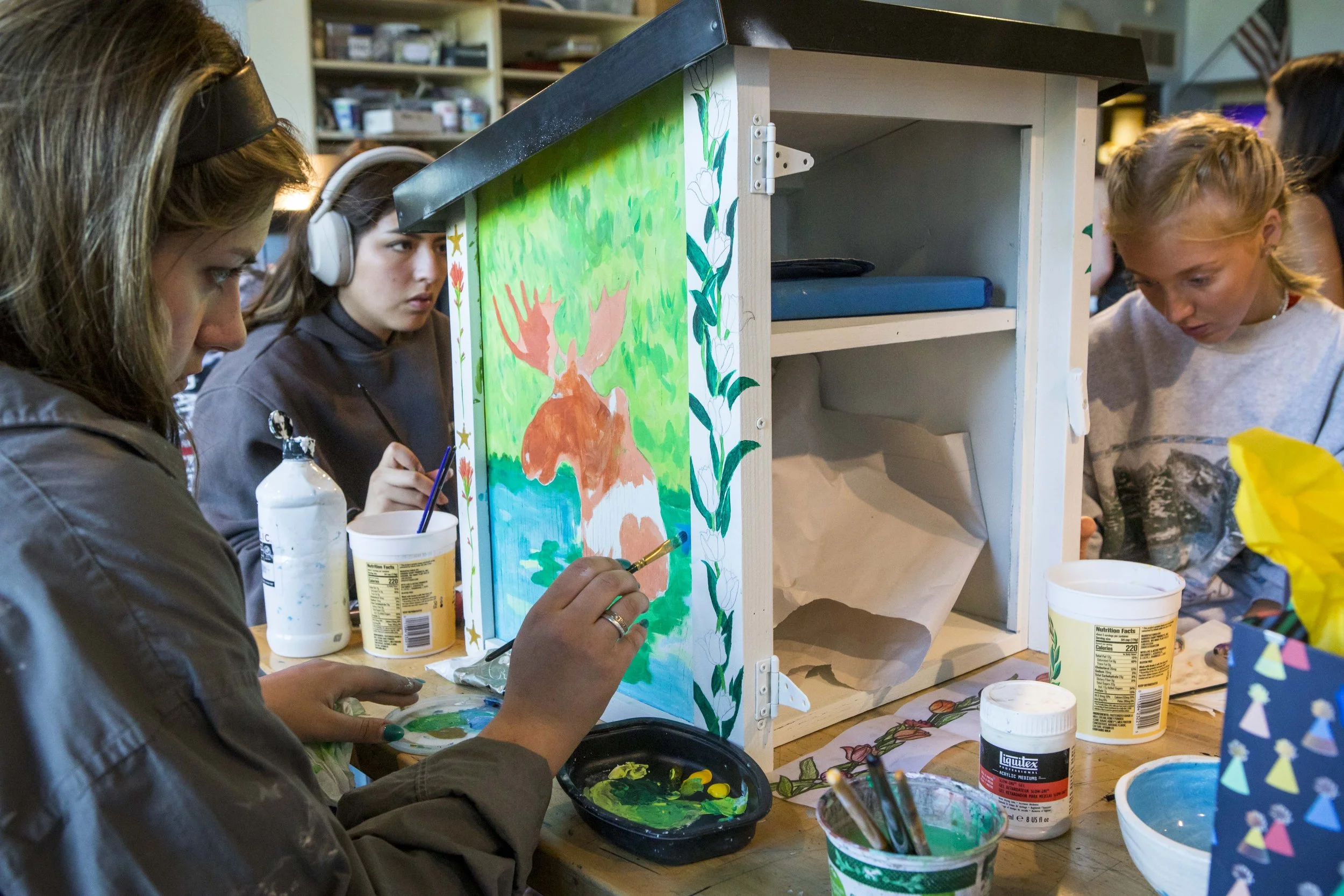 Young girls painting a colorful scene of a deer and trees on a wooden box at a table, surrounded by art supplies in a classroom or art space.