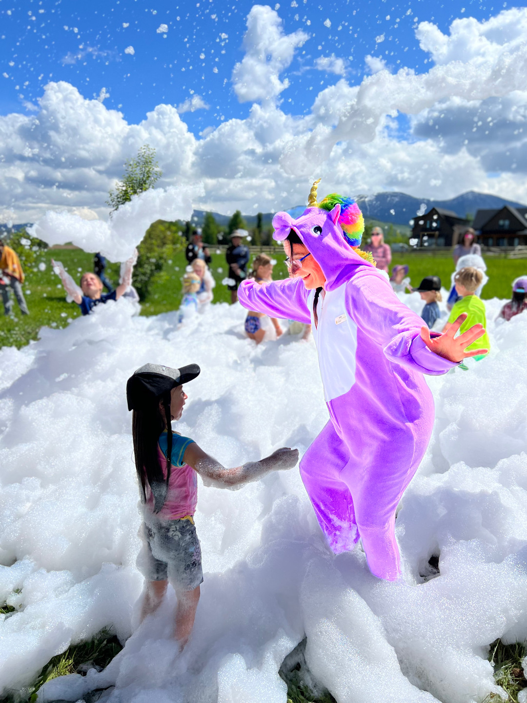 Person dressed in a purple unicorn onesie playing with children in foam bubbles on a grassy field on a sunny day.