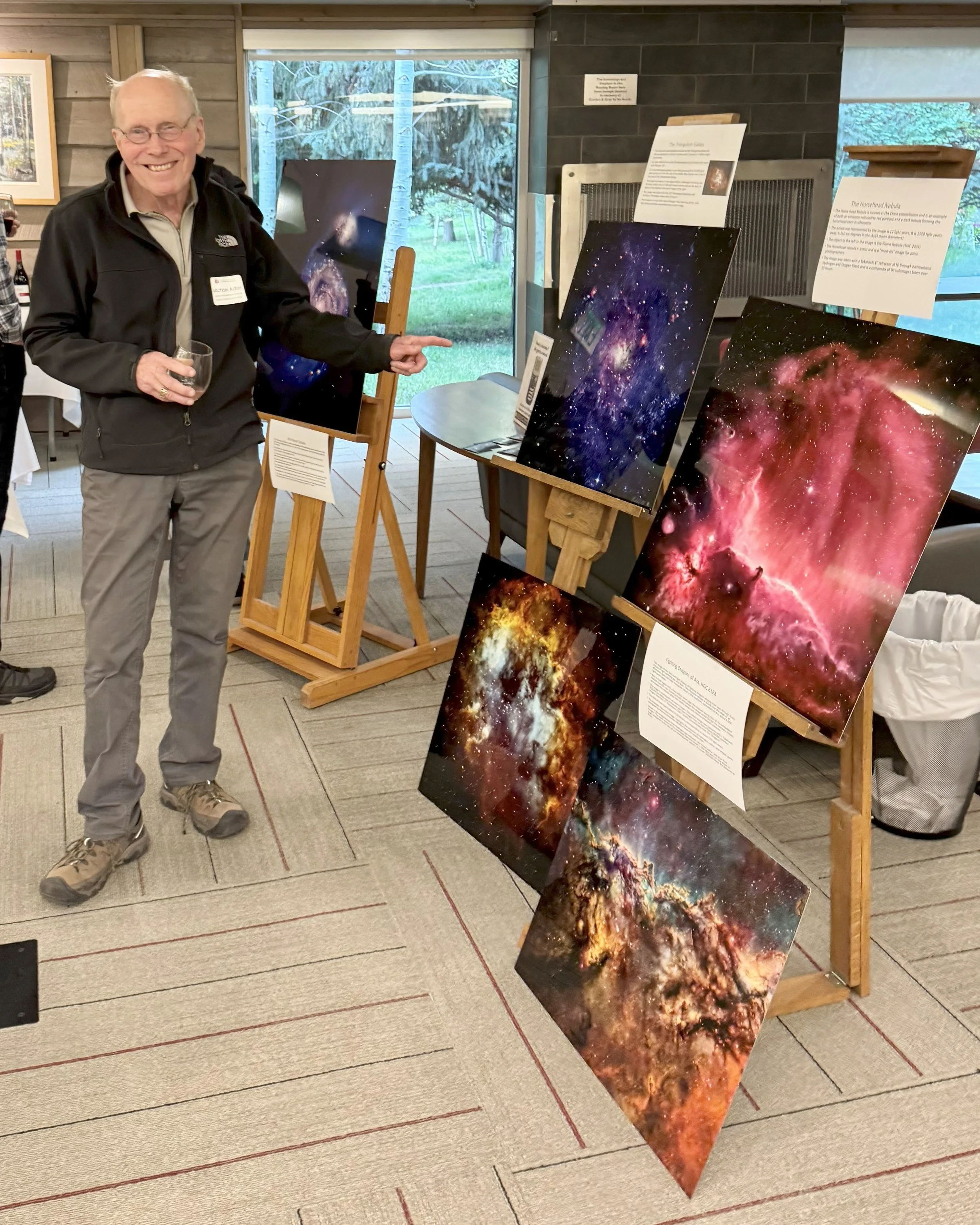 A man holding a glass of wine points at space-themed artwork display in an indoor setting with large windows showing trees outside. The artwork includes images of nebulae and galaxies on easels with informational signs.