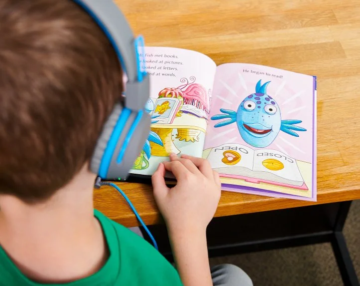 Child with blue headphones reading a colorful children's book at a wooden table