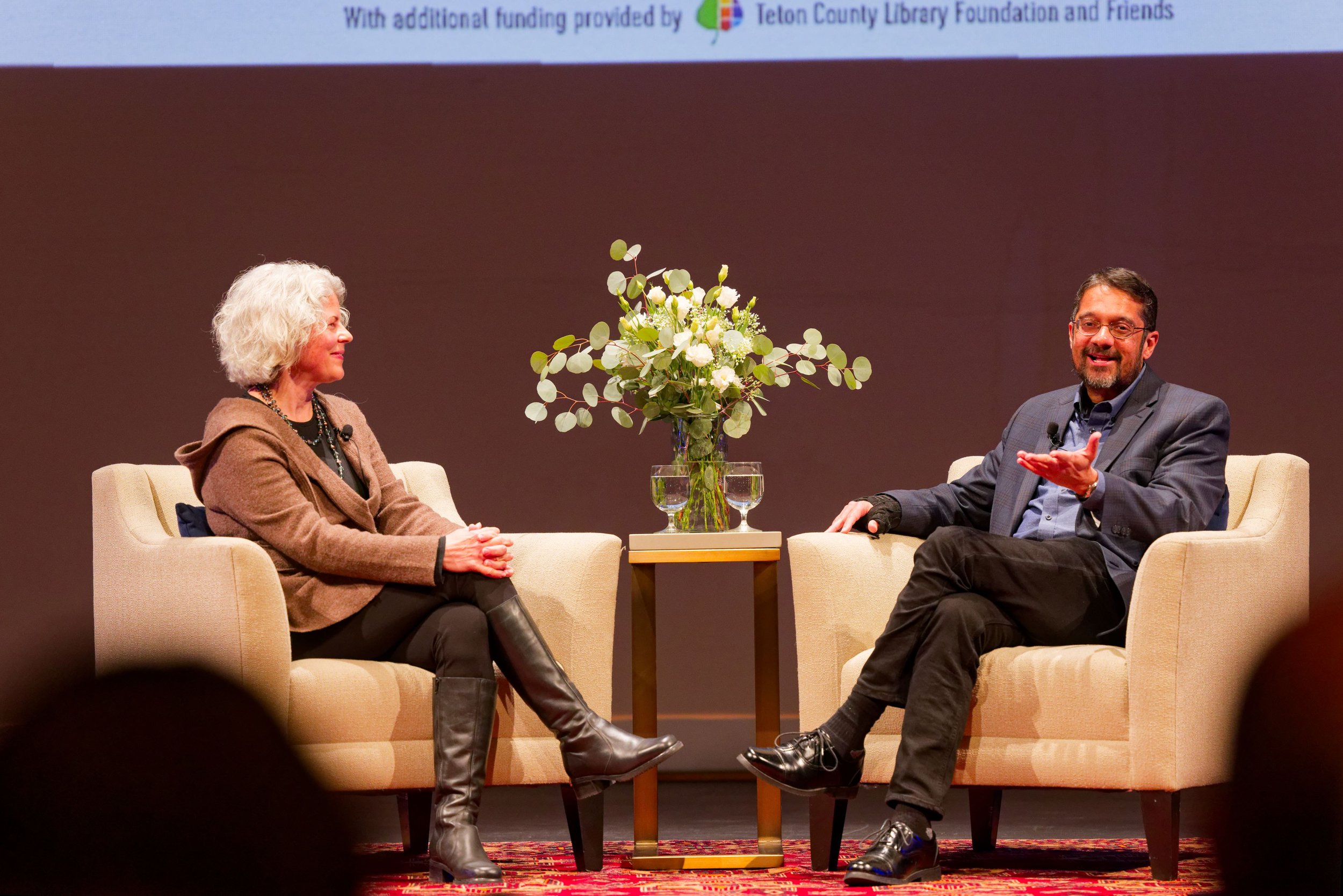 A woman and a man sit in armchairs on a stage, engaged in conversation. A small table with a glass vase of white roses and two glasses of water is between them. The woman has gray hair, and wears a brown blazer, black pants, and tall black boots. The man has dark hair, glasses, and wears a gray blazer, jeans, and black shoes.