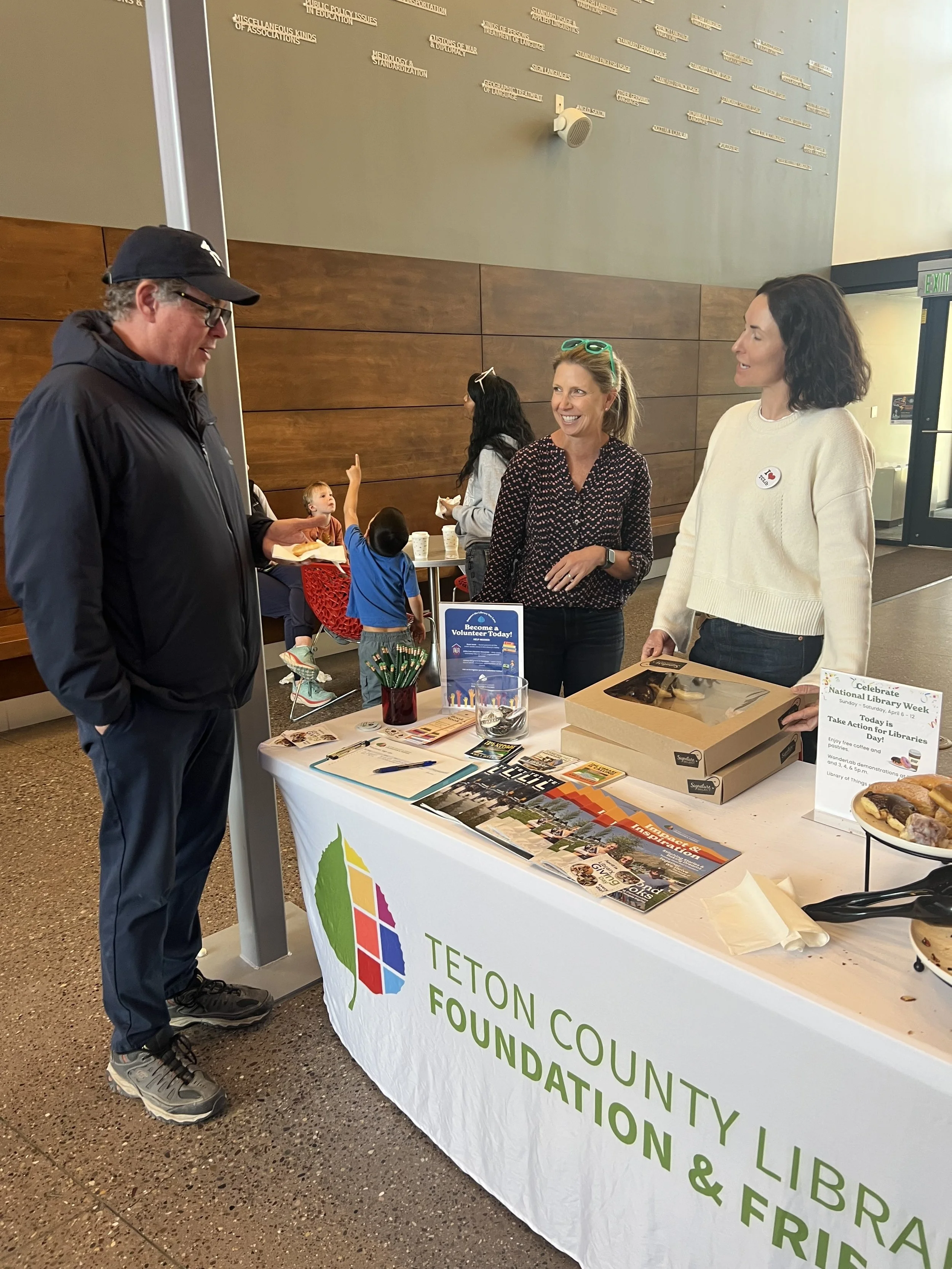 A person talking to two women behind a table at a Teton County Library event promoting library programs and volunteering, with children in the background.