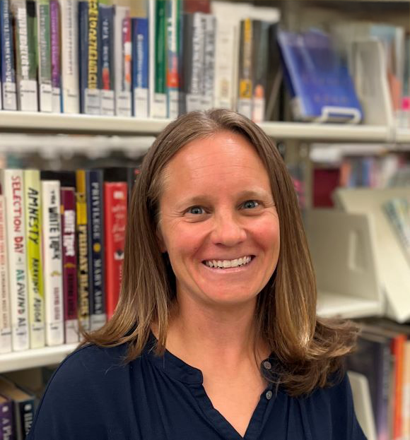 A smiling woman with long brown hair, wearing a navy blue top, standing in front of a bookshelf filled with books in a library.