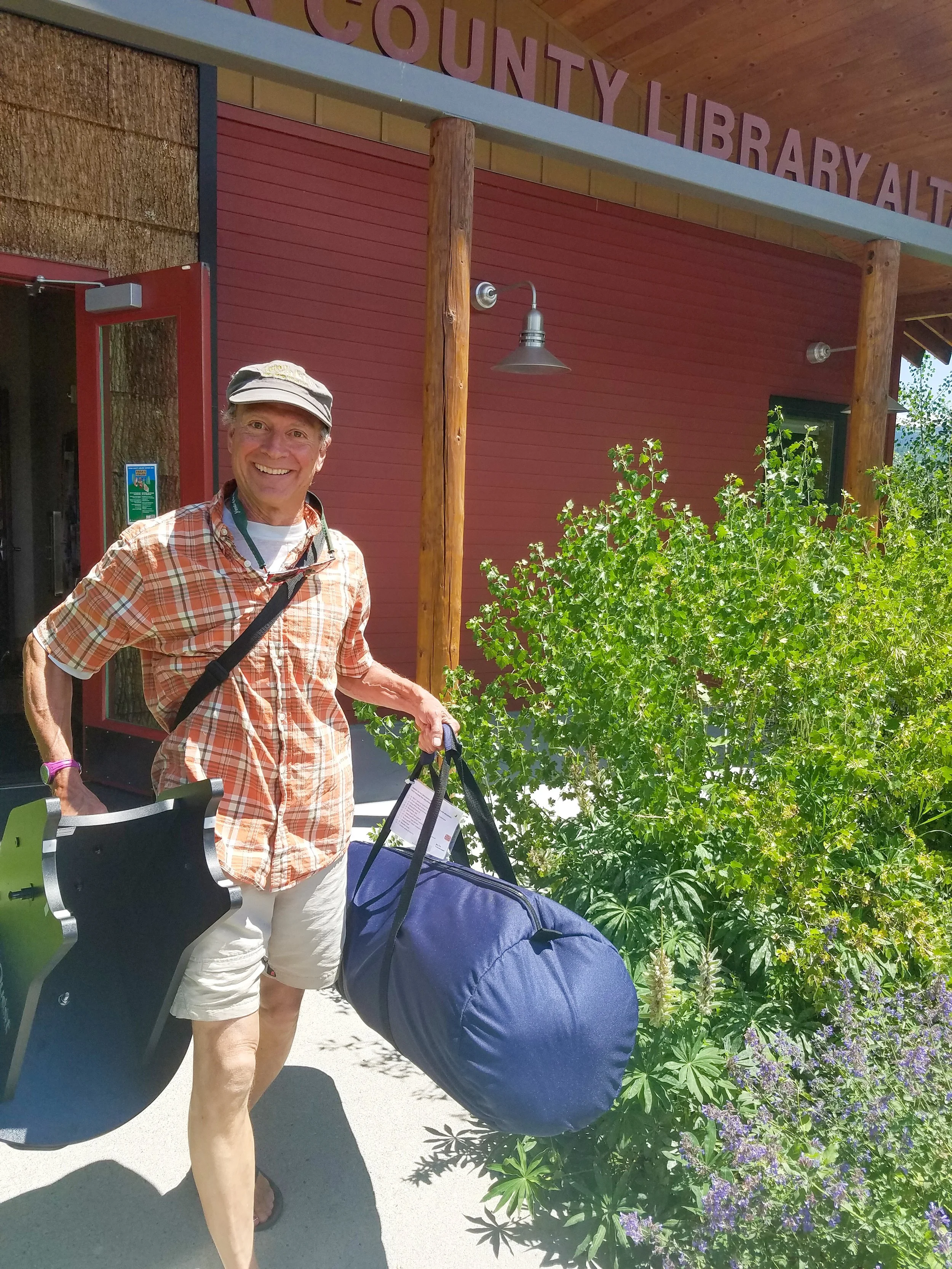 A smiling man with a plaid shirt, shorts, and a hat standing outside a county library, holding a blue duffel bag and a black folding chair, with greenery and purple flowers nearby.