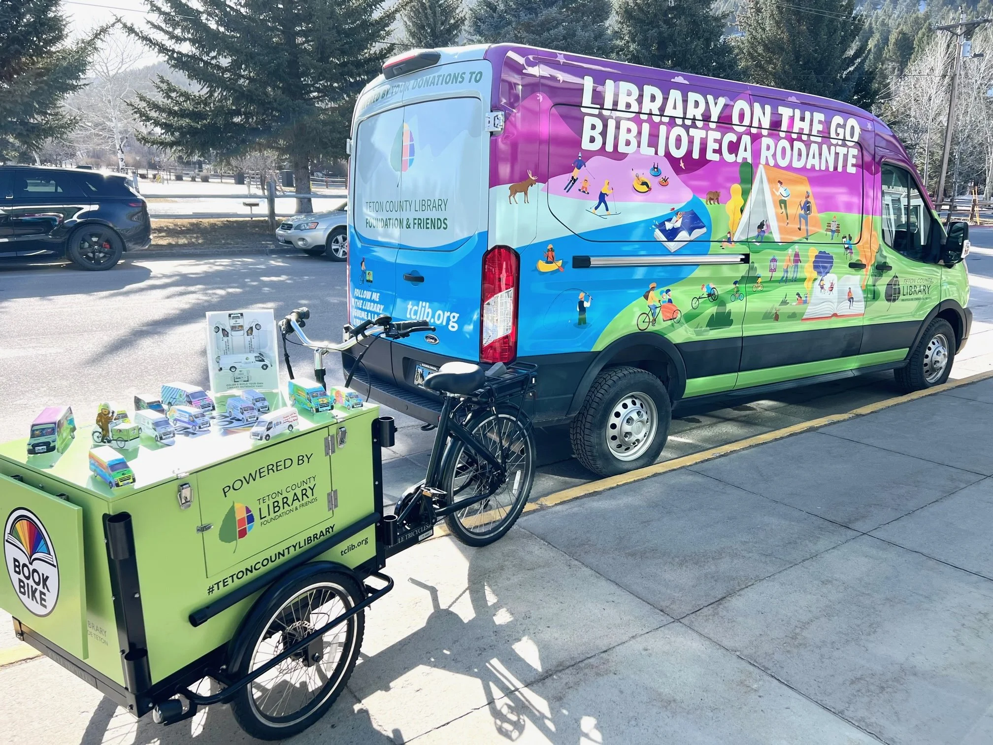 A colorful library on the go vehicle and a green book bike mounted with small toy buses and a yellow toy figure, parked on a sidewalk beside a street with parked cars and leafless trees in the background.