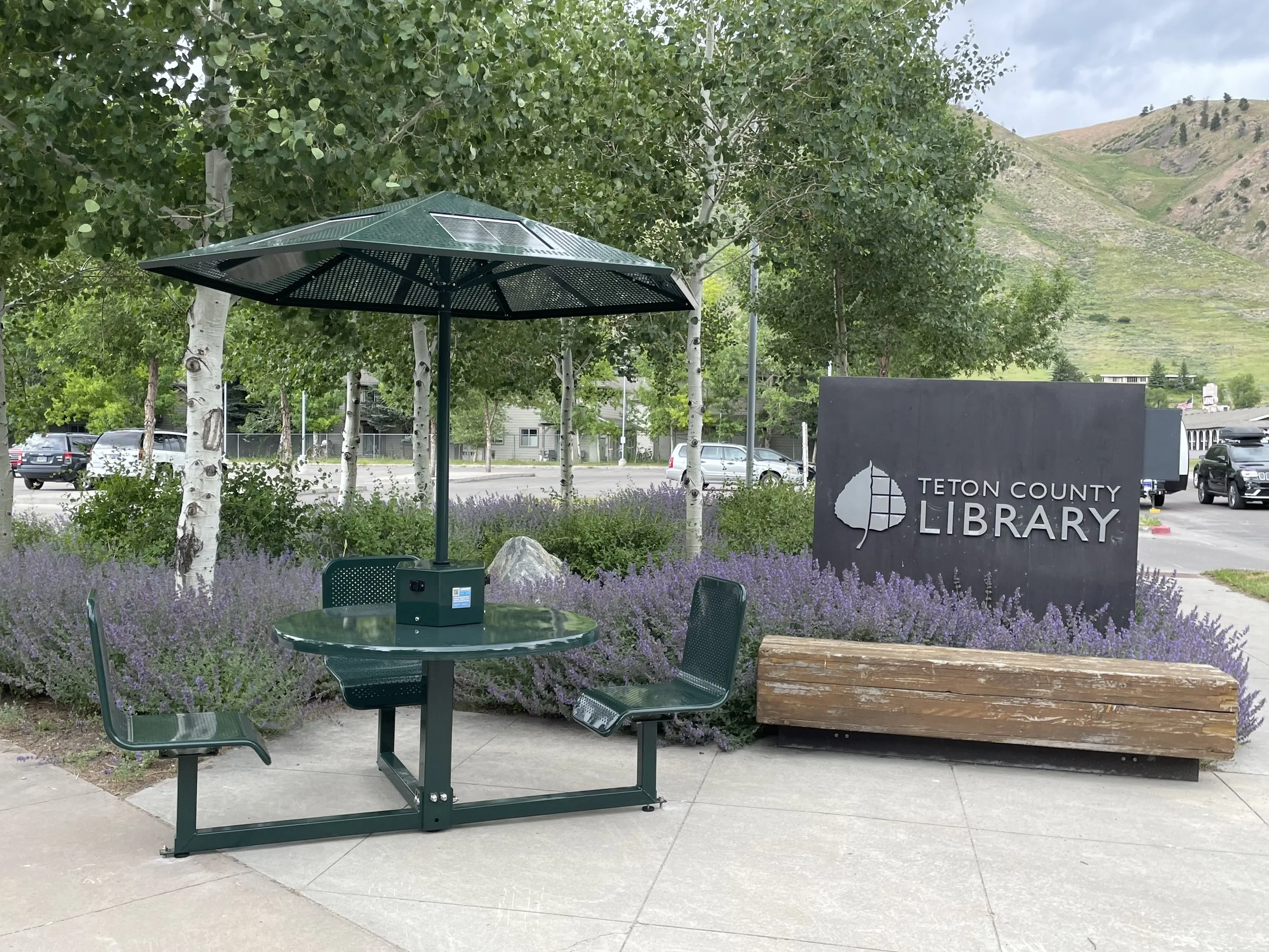 Outdoor seating area with a round green table, three green chairs, a large green umbrella, and a sign for Teton County Library. The area is surrounded by purple flowering plants and green trees, with mountains visible in the background.