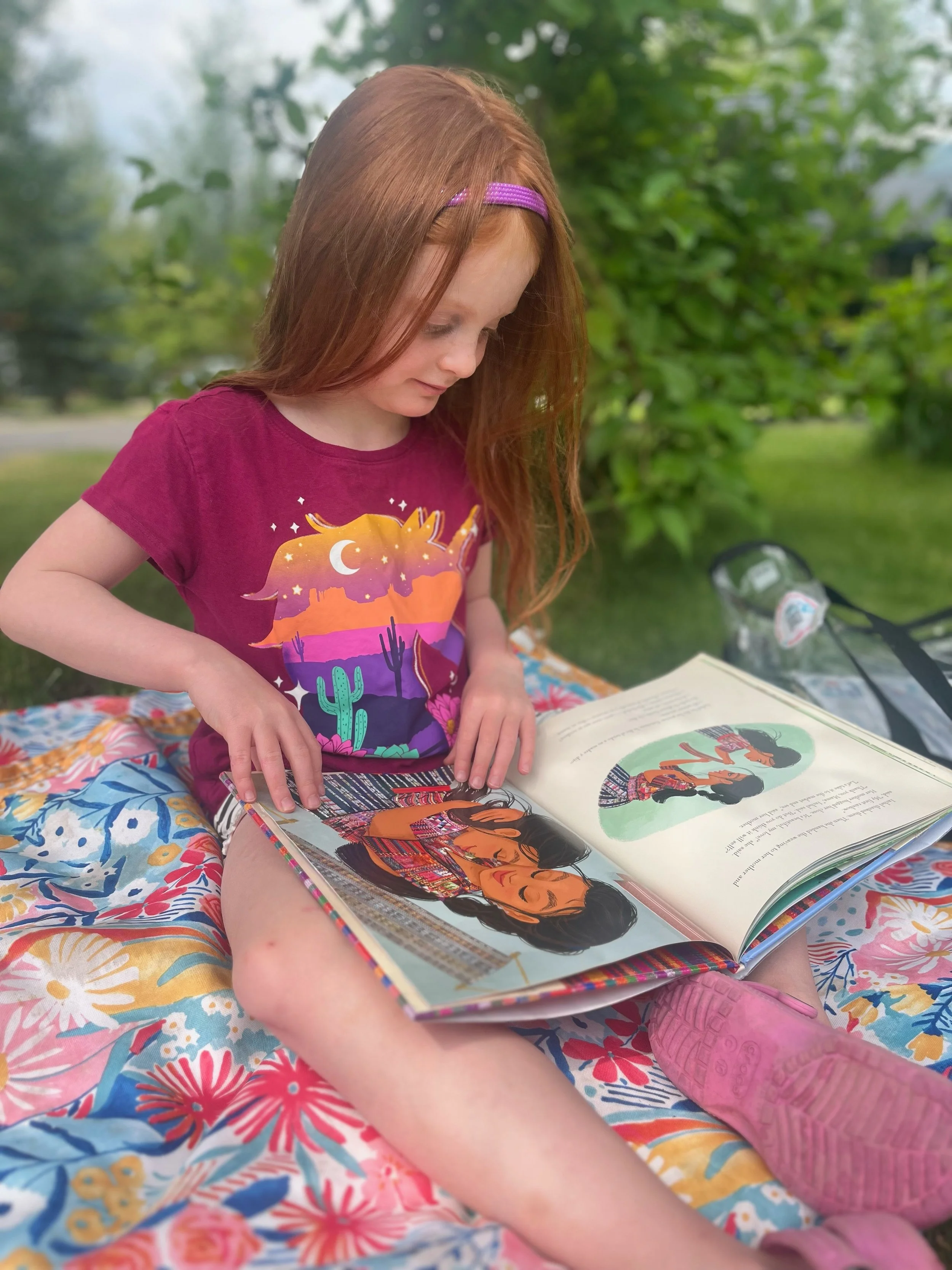 A young girl with red hair, wearing a purple T-shirt with a desert scene and cacti, sits outdoors on a floral blanket reading a colorful picture book. She has a purple headband and pink shoes, with trees and greenery in the background.