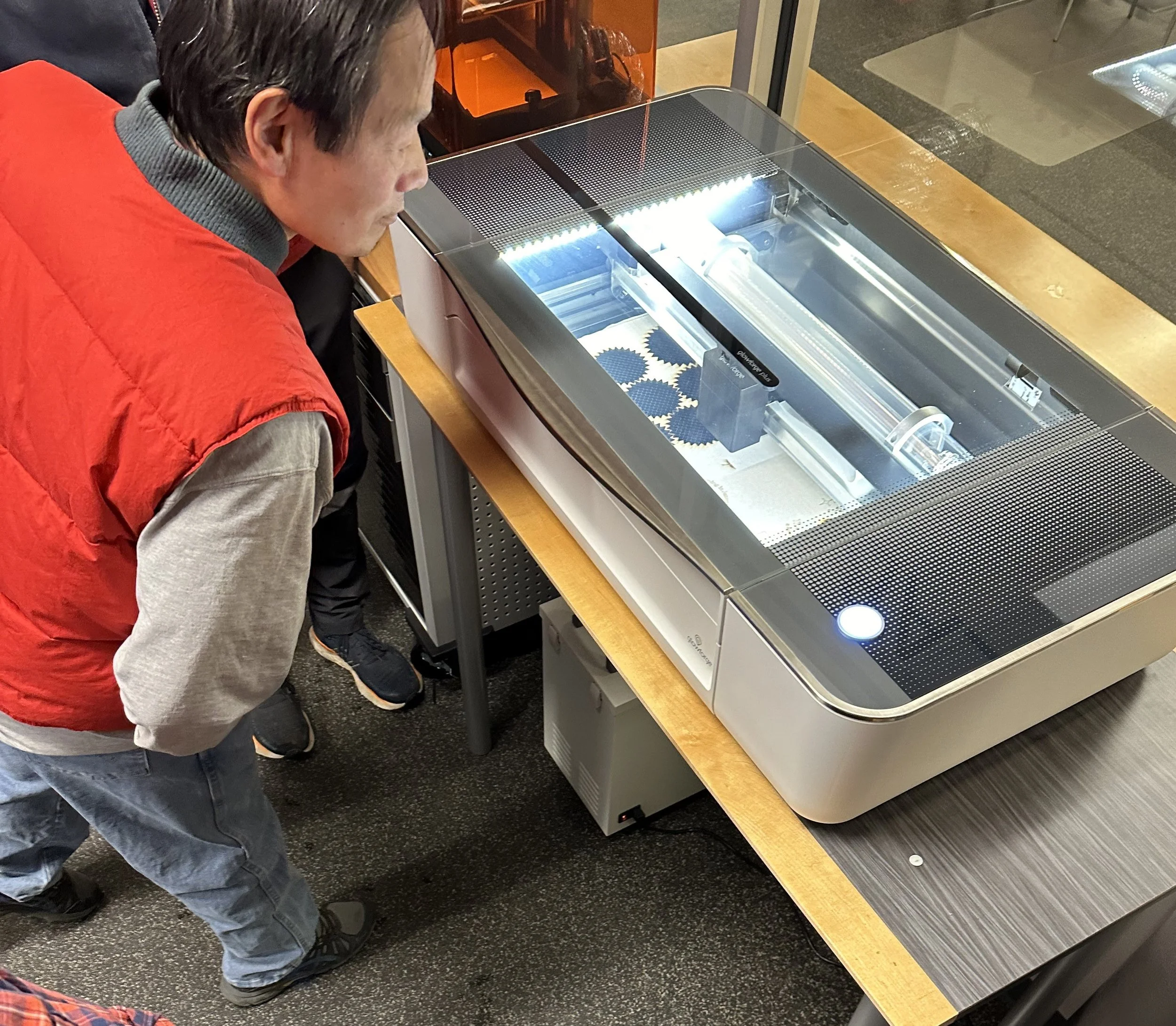 A person looking at a tabletop 3D printer with a glass cover, which is printing small gear-shaped objects.