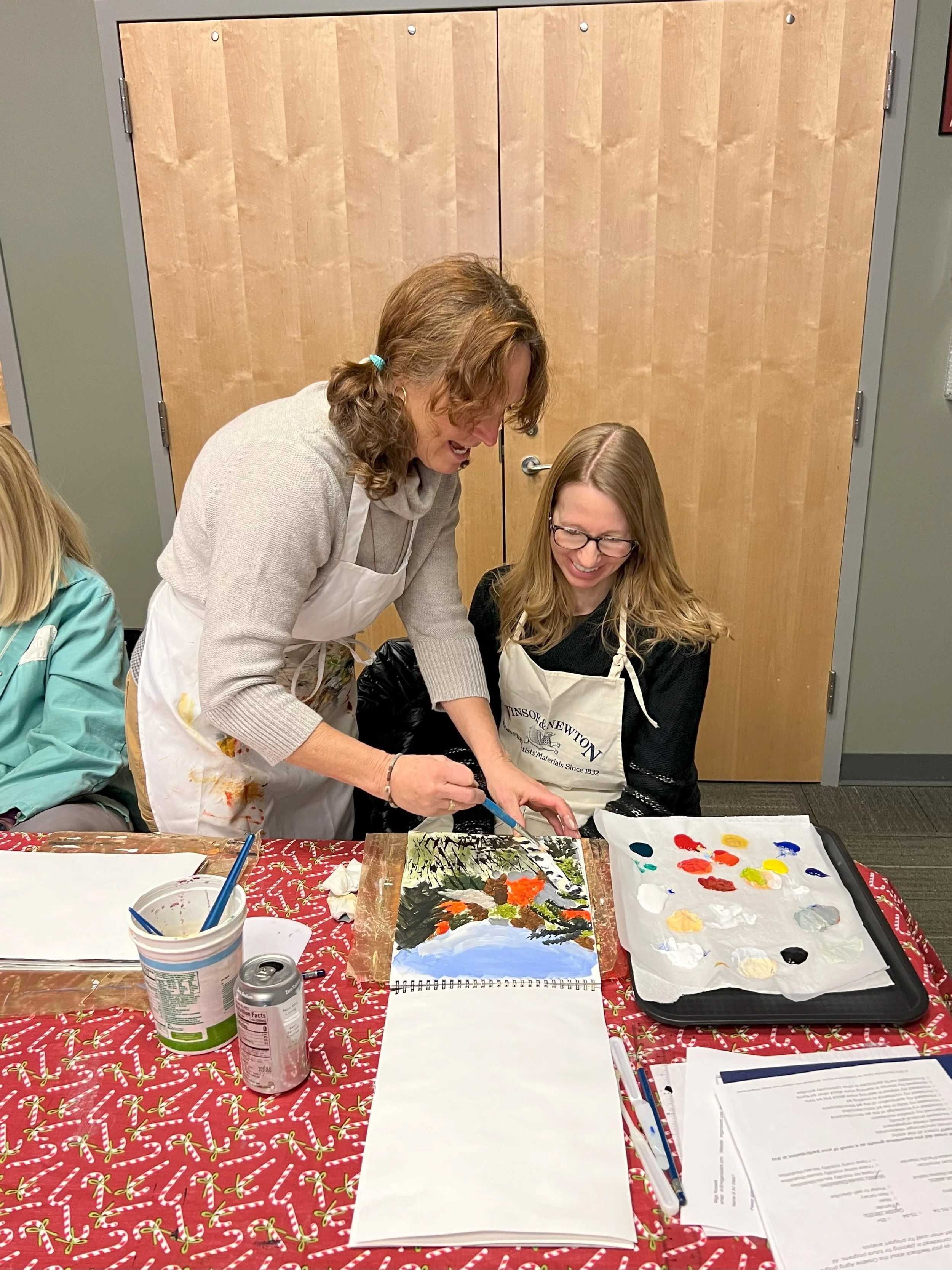 Two women are painting together at a table covered with a red holiday themed tablecloth. One woman, standing and wearing a white apron, is guiding the other woman, seated and wearing glasses and a similar apron. There are paint supplies, paper, and a painted artwork on the table.