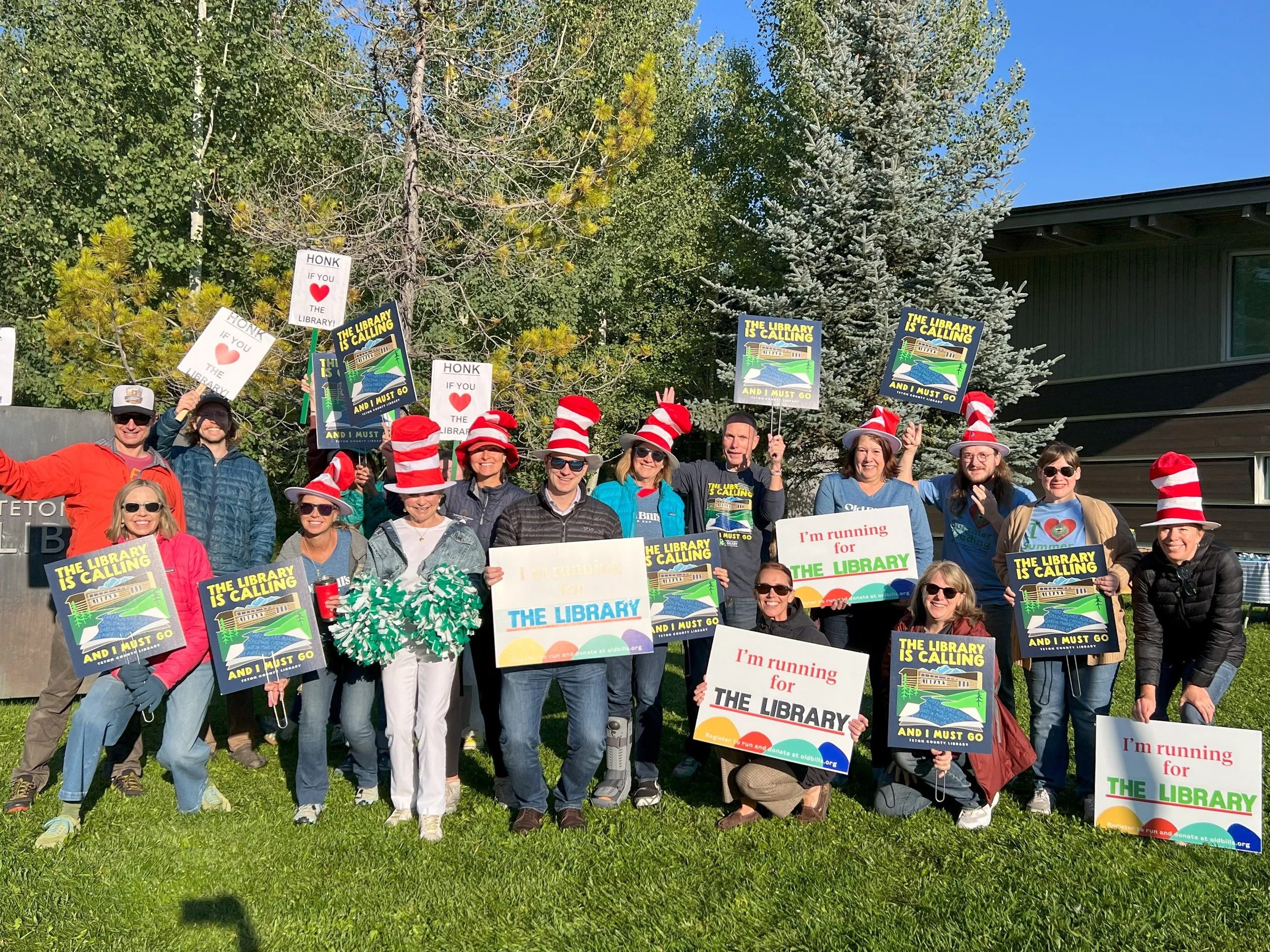 Group of people holding signs that promote the library, some wearing Dr. Seuss-style red and white striped hats, outdoors on a sunny day with trees and a house in the background.