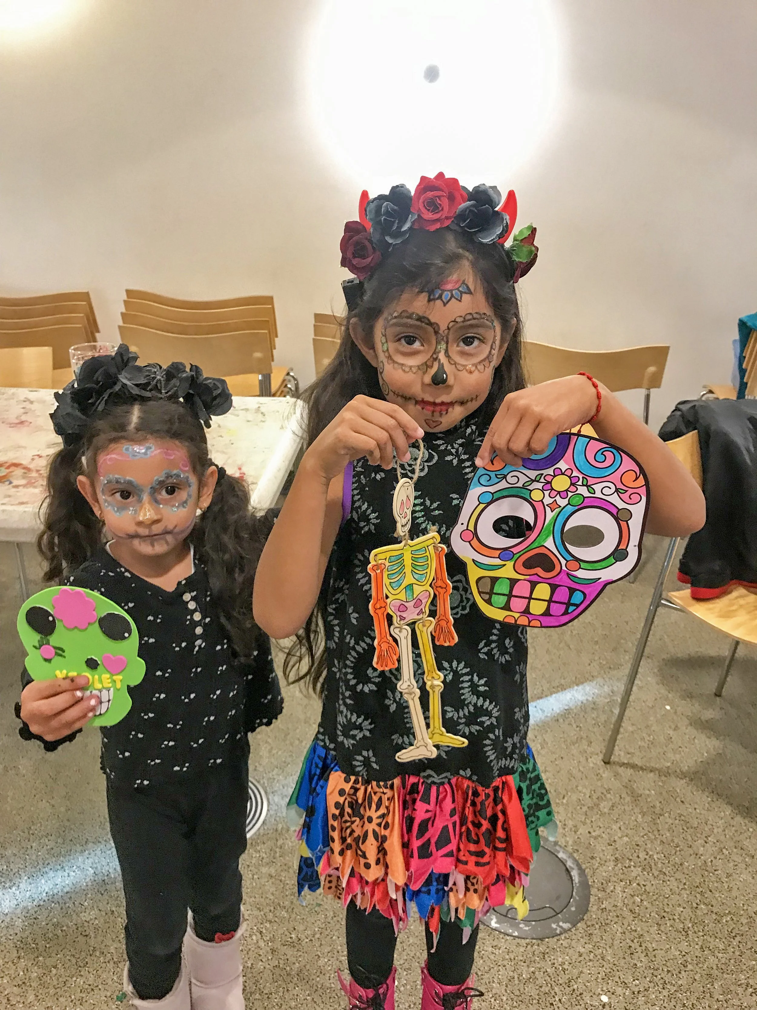 Two young girls in traditional Mexican sugar skull face paint, holding colorful skull and skeleton decorations, celebrating Dia de los Muertos.