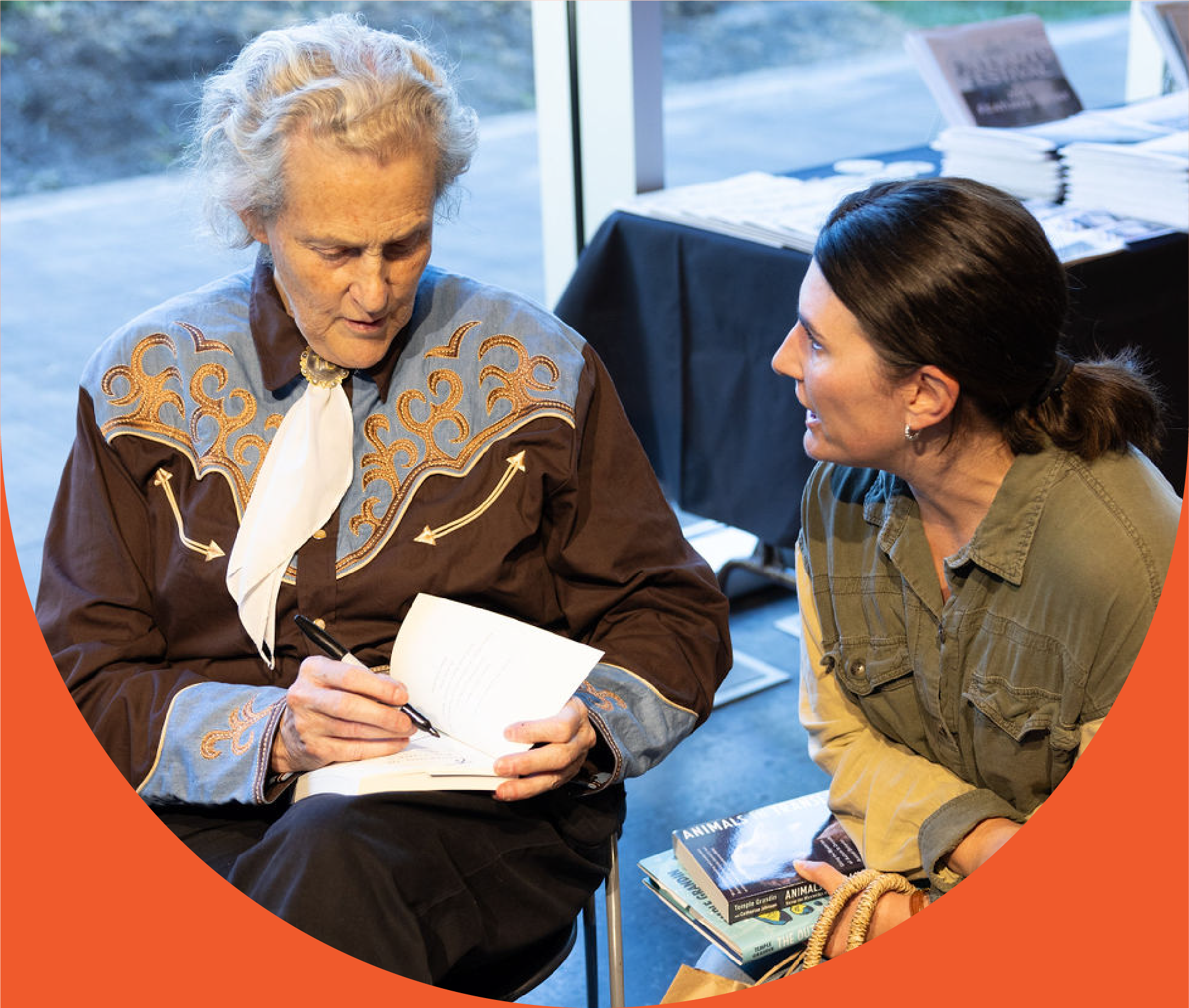 Two women sitting and engaging in a conversation, one older with gray hair signing a book, and a younger woman with dark hair looking at her. They are in an indoor space with a window and a table with newspapers and books in the background.