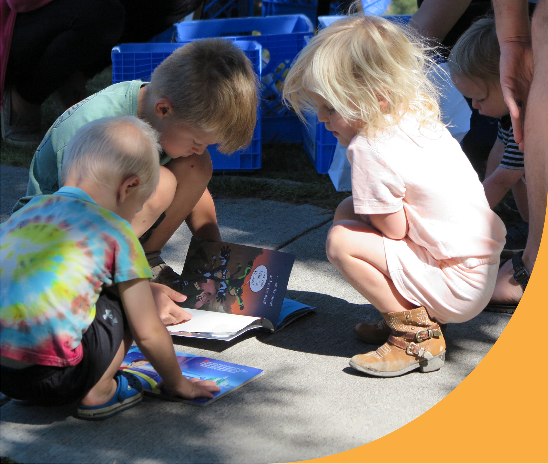 Children kneeling and sitting on the ground, looking at open picture books during an outdoor activity.