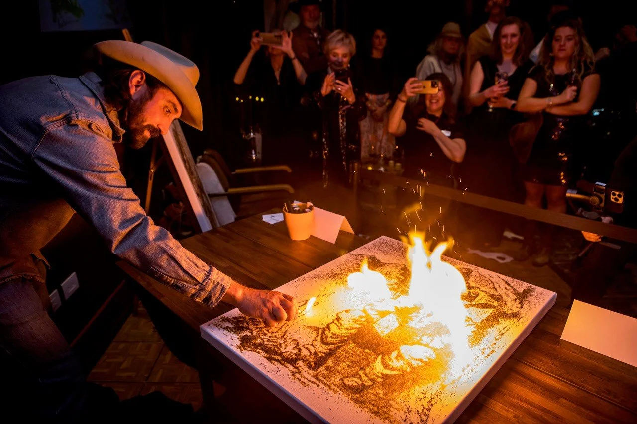 A man in a cowboy hat and denim shirt creates a fire art on a canvas with a torch, in front of an audience taking photos and videos.