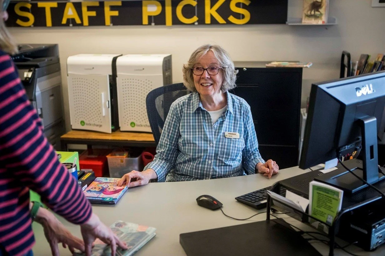 A cheerful older woman with blond hair and glasses sitting at a desk, talking to a person wearing a striped shirt. The woman has a name tag and is in a classroom or office environment with a black sign reading 'STAFF PICKS' on the wall behind her, along with books and office supplies.