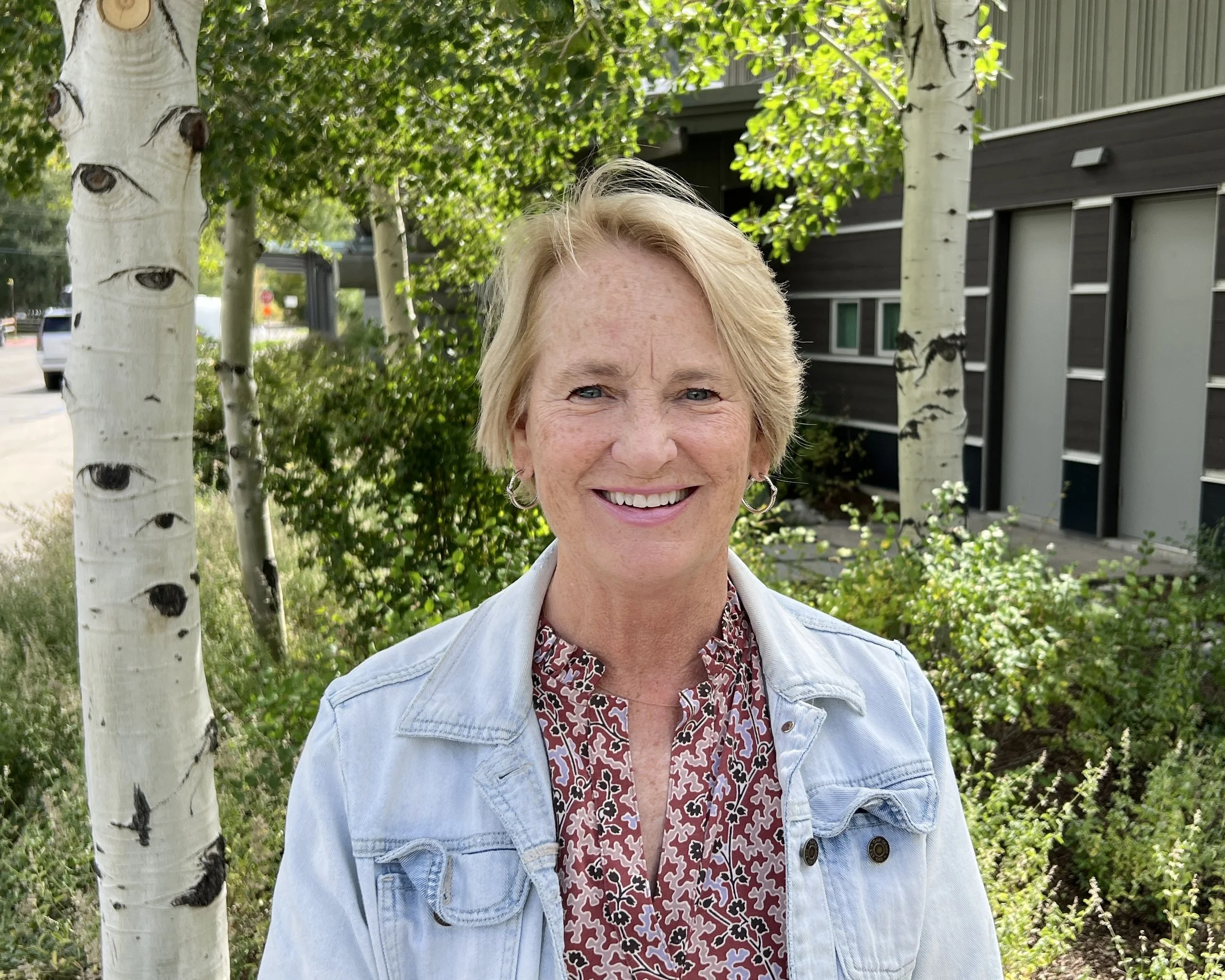 Smiling woman with short blonde hair, wearing a red patterned blouse and light blue denim jacket, standing outdoors in front of a tree with white bark and green leaves, and a modern building in the background.