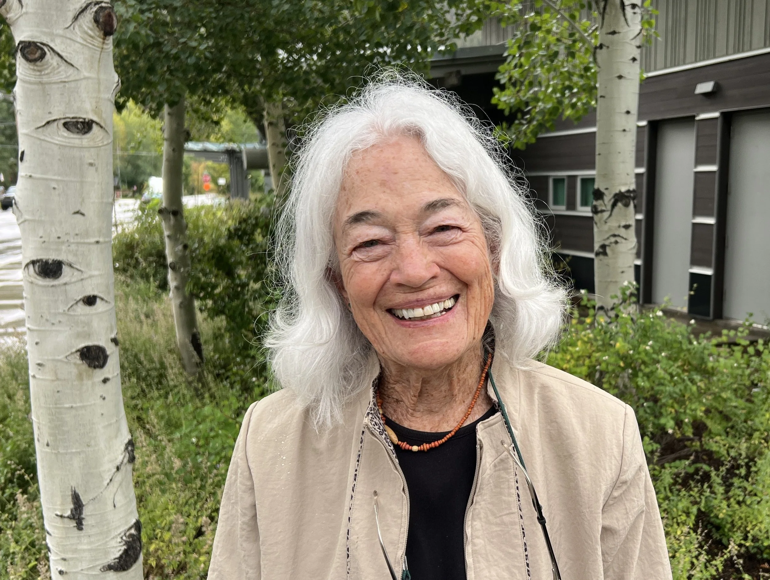 Smiling elderly woman with white hair standing outdoors near trees and a modern building.