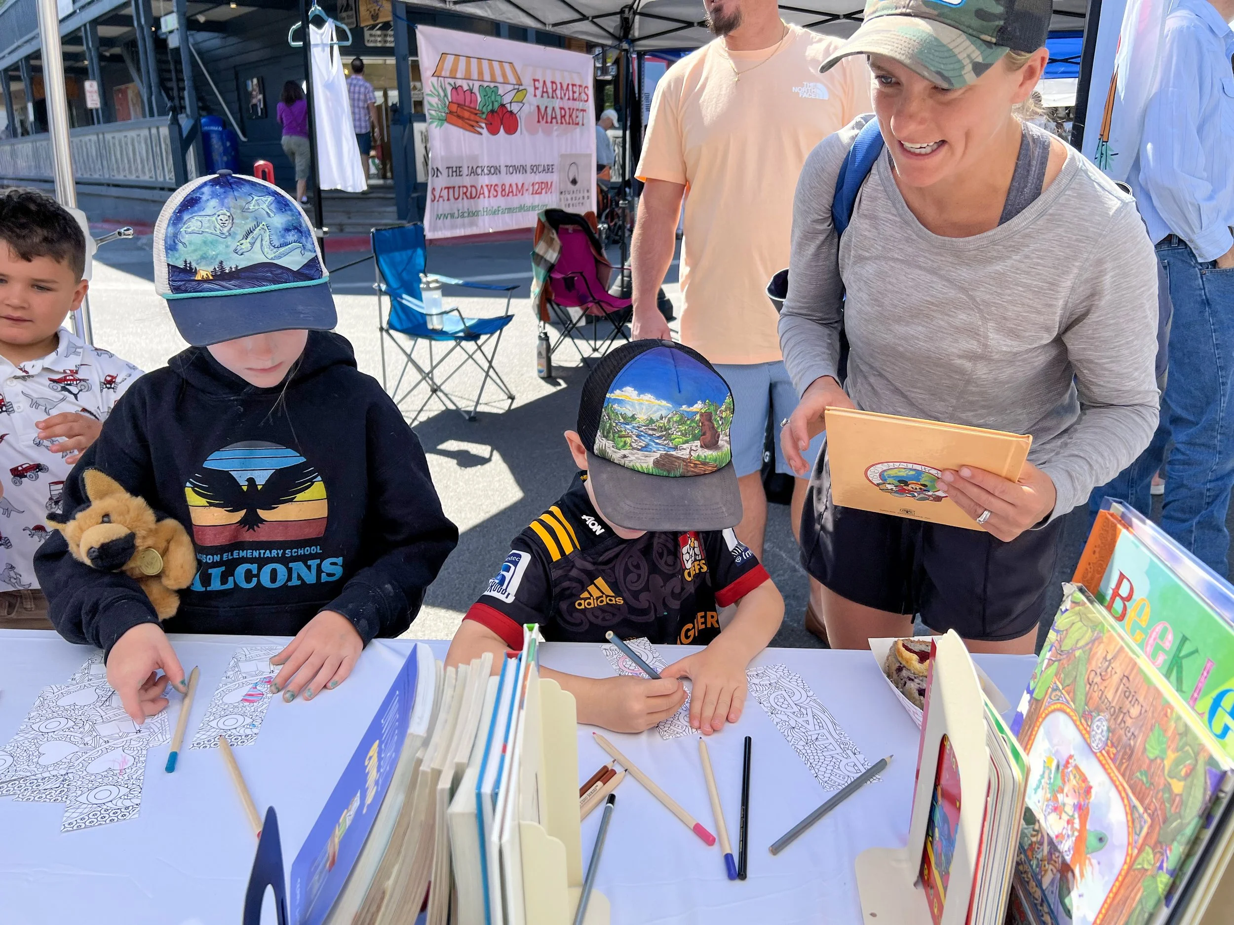 A woman is smiling and talking to two children at a table with coloring books and markers at an outdoor farmers market. The children are coloring pictures, one holding a plush toy and the other wearing a cap with a mountain scene. There are more people and market tents in the background.