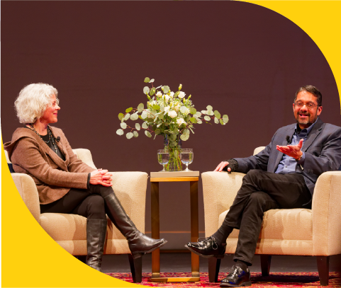 A woman and a man sitting in beige armchairs on a stage, having a conversation. A small table with a large bouquet of white flowers is between them. The background is a plain brown wall with a yellow corner.