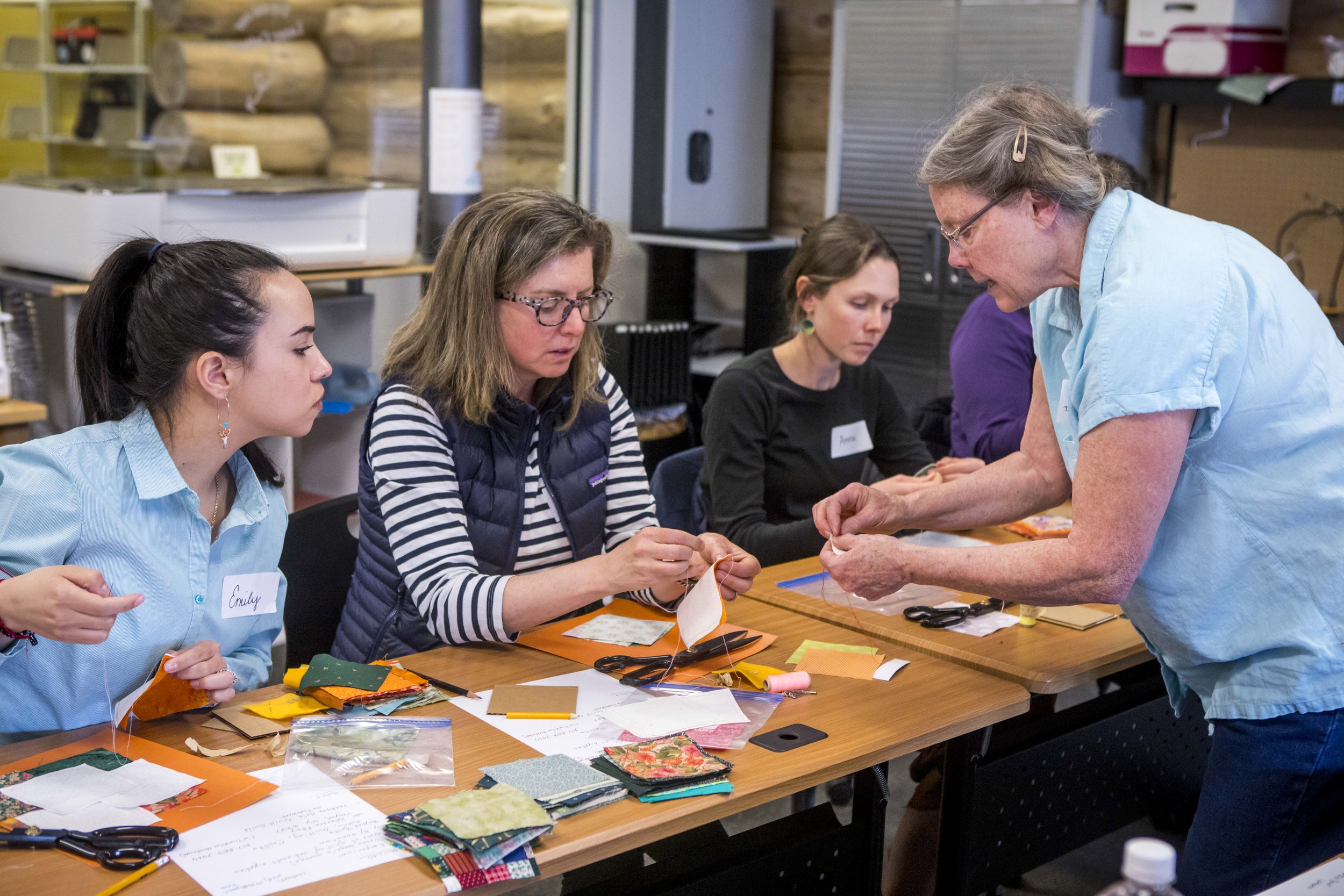 A group of women sitting at a table with fabric pieces, scissors, and sewing supplies, participating in a fabric craft workshop, with an older woman assisting.
