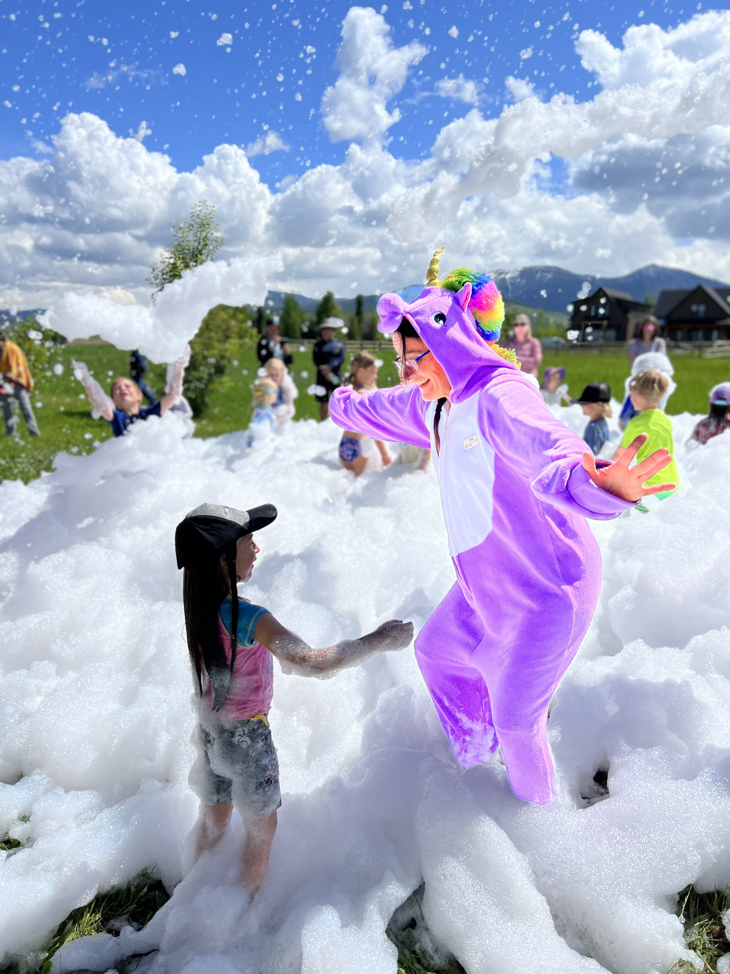 People, including children, playing in a large foam party outdoors on a sunny day with mountains and houses in the background. One person is dressed in a purple unicorn costume with a rainbow mane.