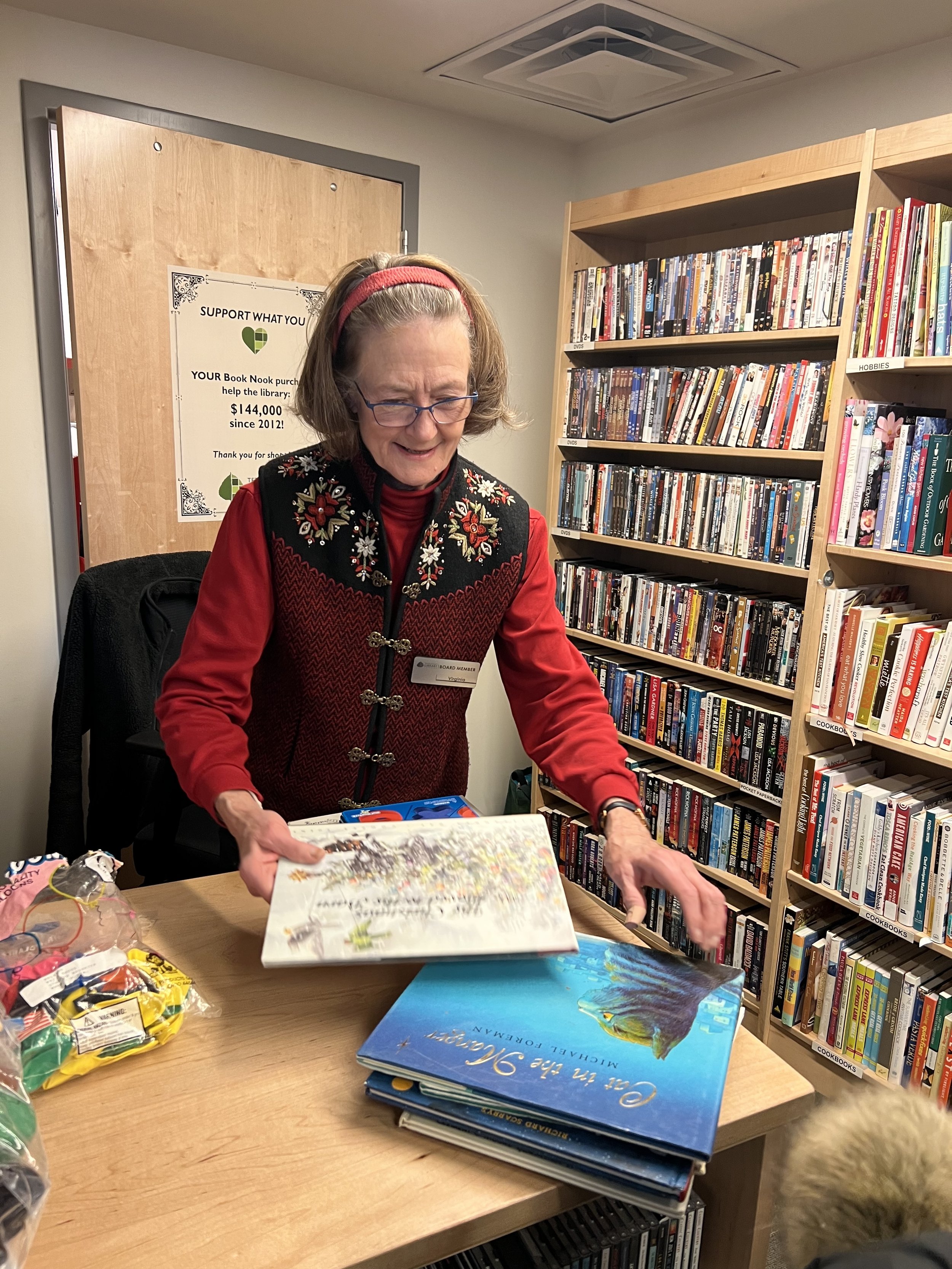 A woman with glasses, wearing a red and black embroidered vest, is smiling and looking at books on a table in a library. There are several books and bags of snacks on the table, with bookshelves filled with DVDs and books in the background.