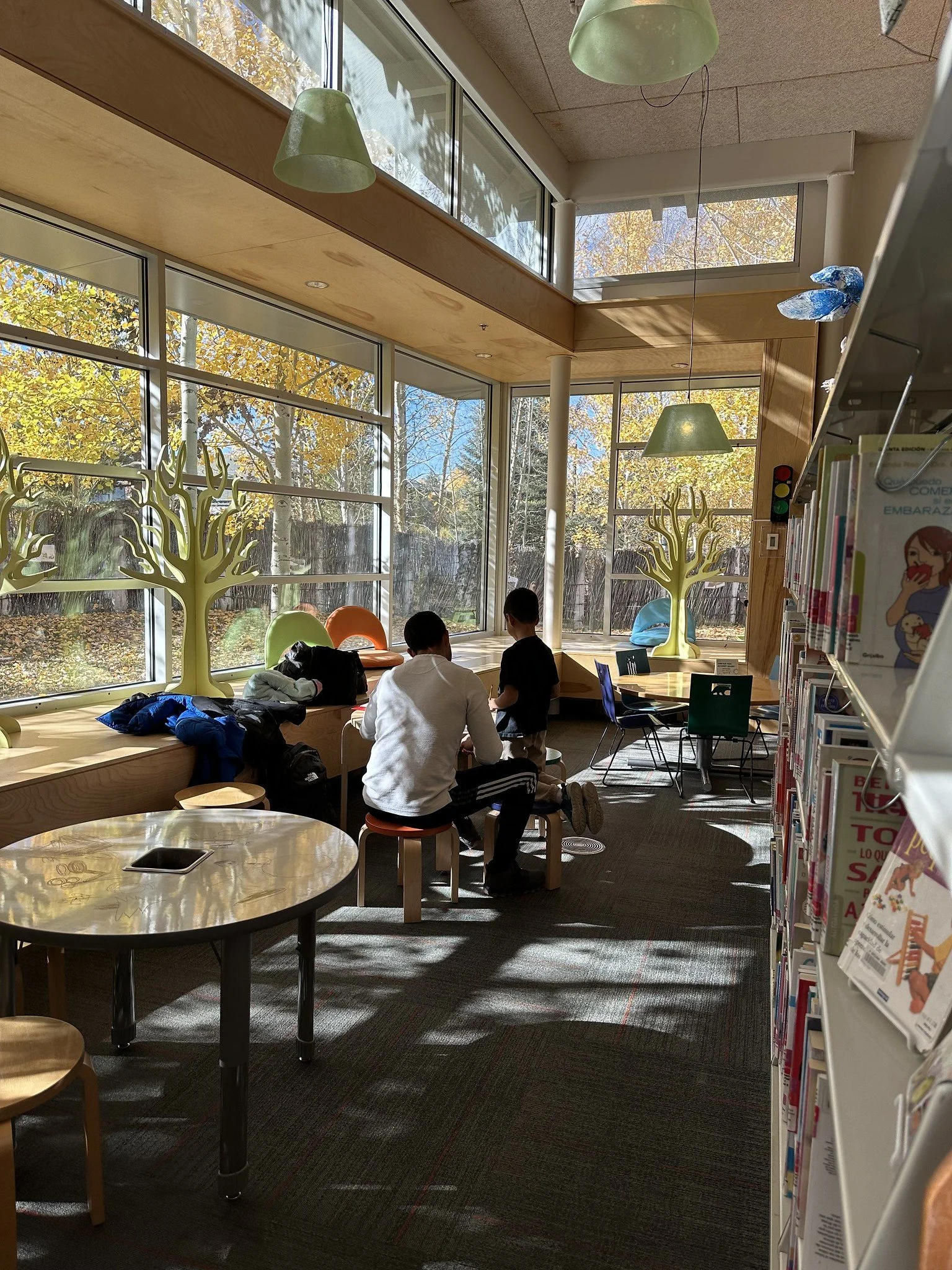 A brightly lit library corner with large windows showing fall foliage outside. There are two decorative yellow trees on the windowsills, a man and a boy sitting and talking at a small table, with backpacks nearby. Sustainable green hanging lamps illuminate the space, and a colorful child’s chair and bookshelves with magazines are visible.