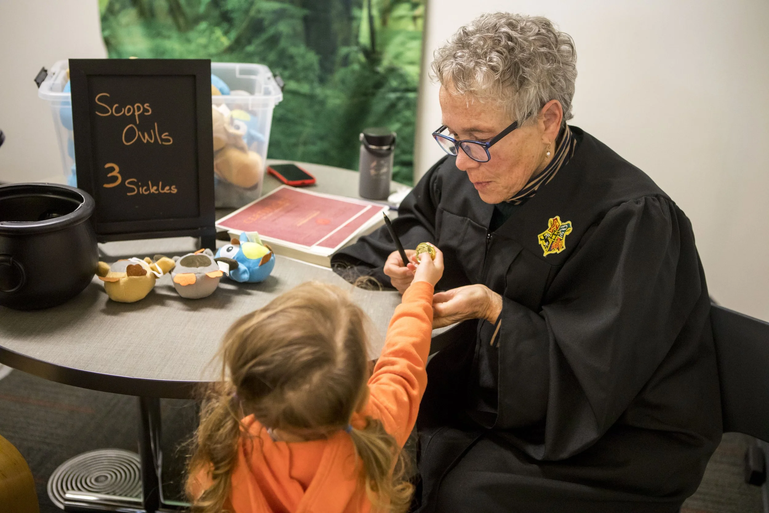 An woman dressed in a black witch's robe is sitting at a table, signing an item for a young girl wearing an orange hoodie. The table displays plush toys resembling owls with various colors and accessories.