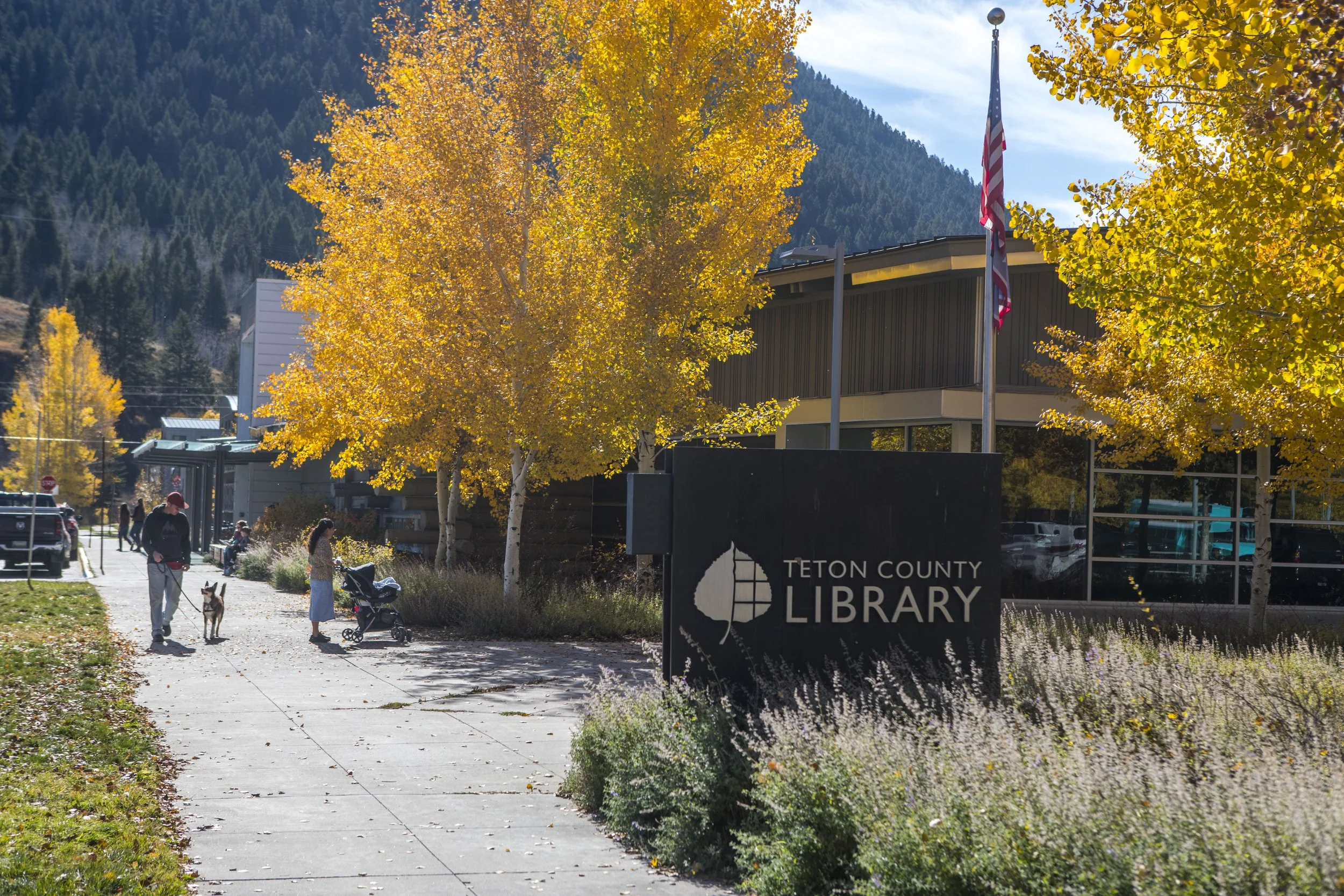 The Teton County Library exterior with a sign, trees with yellow autumn leaves, people walking with a dog and a stroller, and a mountain in the background.