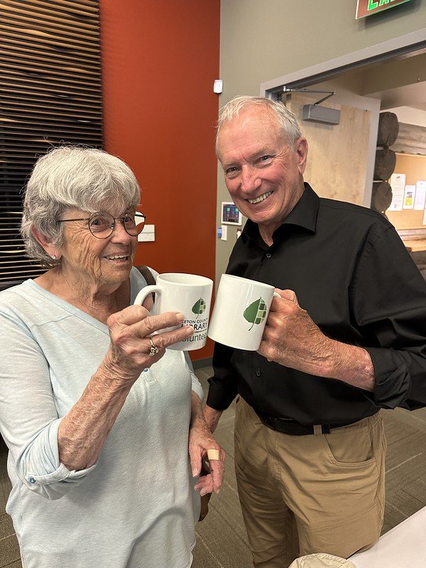 An elderly woman and a middle-aged man smiling and holding white mugs with green leaf logos, indoors in a modern space.