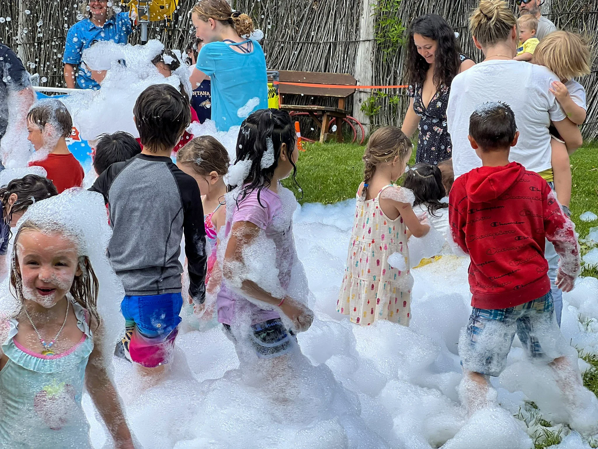 Children and adults playing in a foam party outdoors, with foam on the grass and children enjoying and laughing.