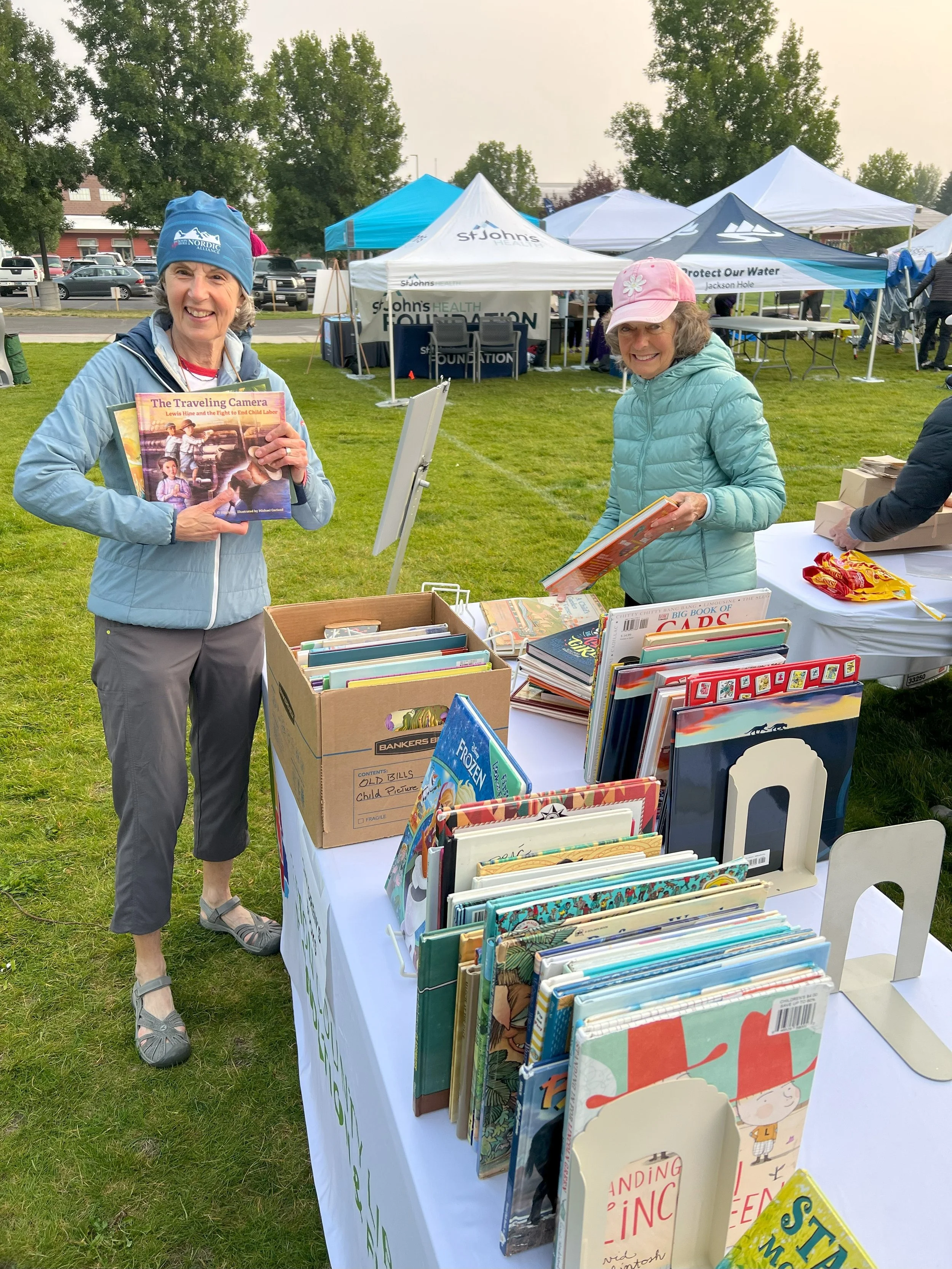 Two women at an outdoor book sale, one holding a book titled 'The Traveling Camera' and the other looking at books on a table with cartoons and children's books, no specific background or location details.