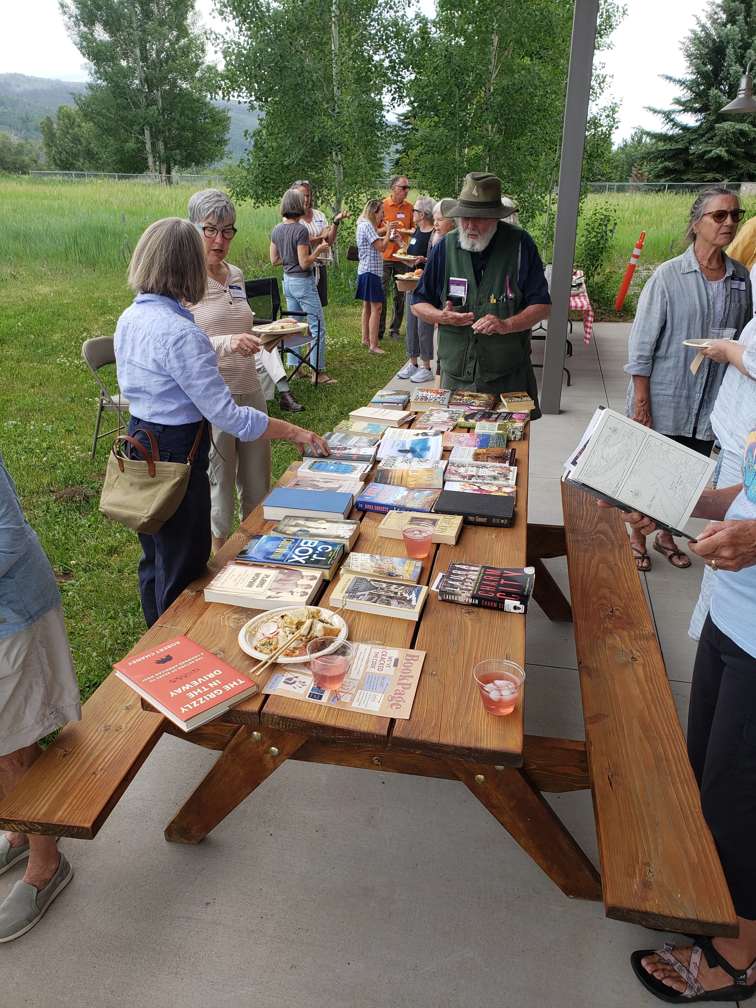 People browsing books on a table during an outdoor book event with green grass and trees in the background.