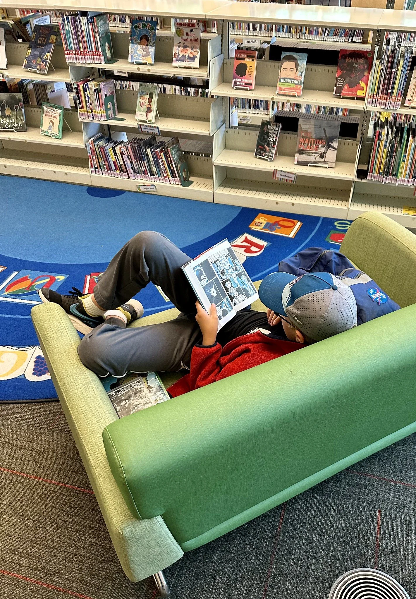A child lying on a green couch and reading a comic book in a library, with bookshelves in the background.