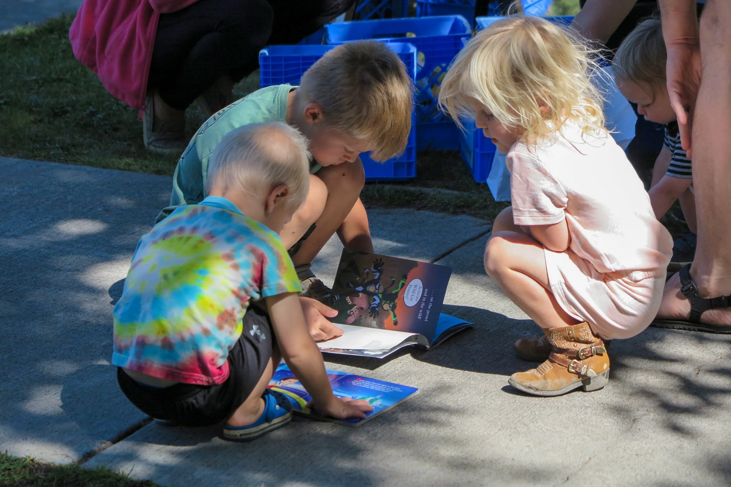 Three young children crouching on a sidewalk, looking at books, with two adults nearby and transparent blue crates in the background.