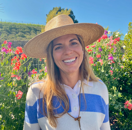 A woman smiling outdoors in a garden with pink and red flowers, wearing a large straw hat and a striped blue and white top.