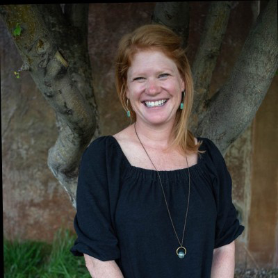 A woman with red hair smiling outdoors, standing in front of a tree.