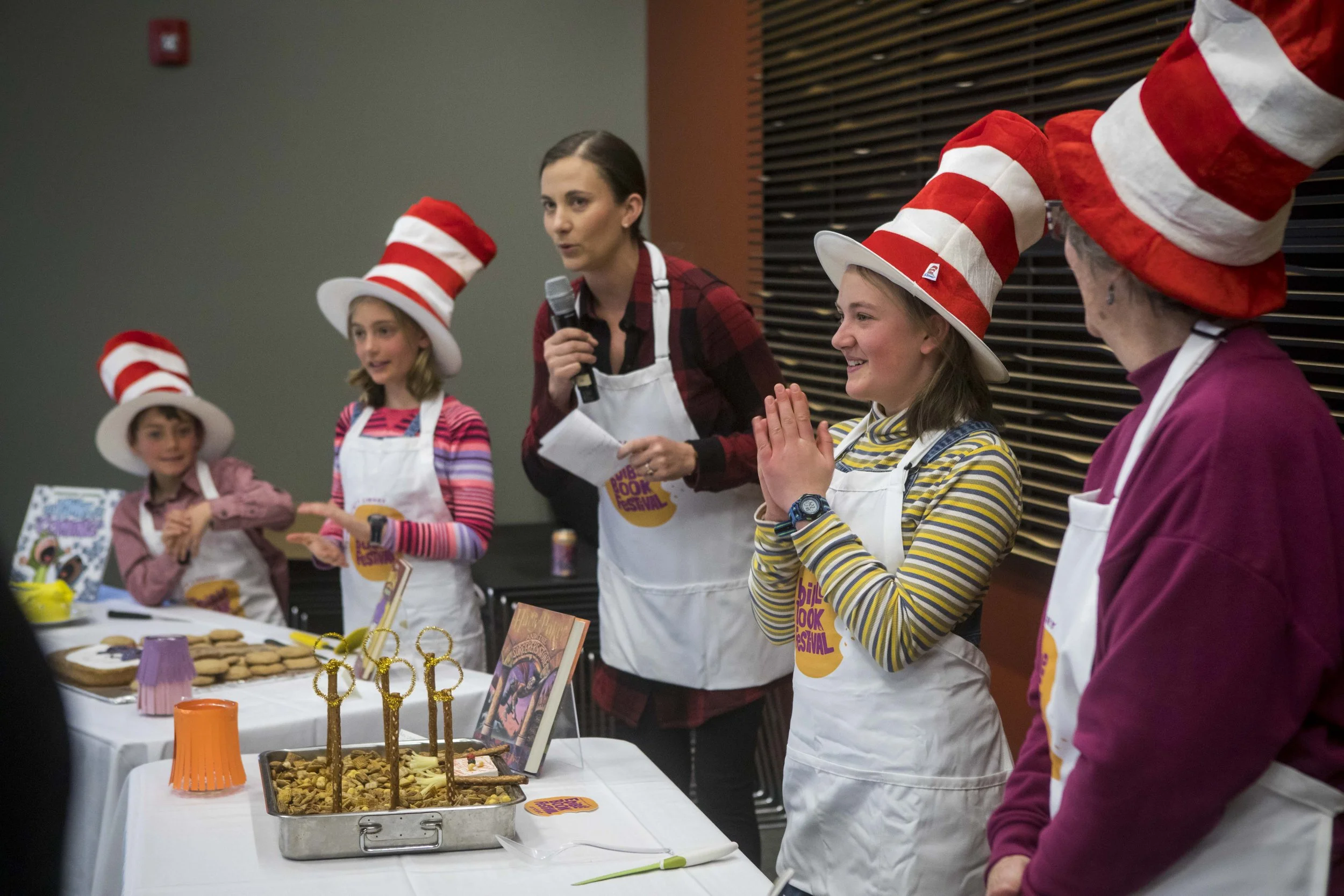Children and an adult at a book festival wearing red and white striped hats, with a table of cookies and treats.