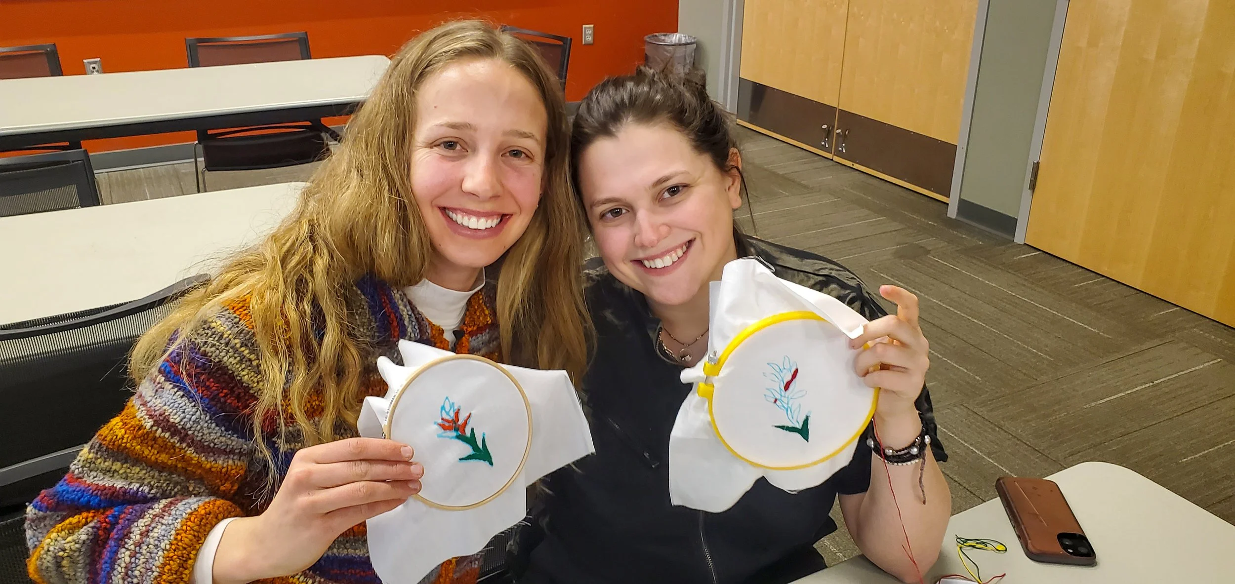 Two young women sitting at a table, smiling and showing embroidered pieces they made, in a room with wooden doors and orange walls.