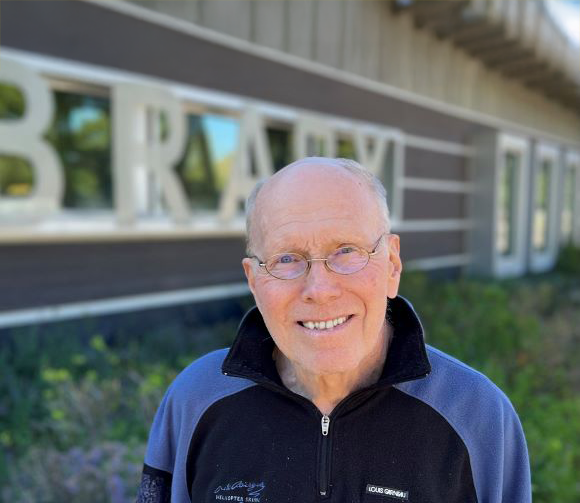 Smiling elderly man with glasses and a blue and black jacket standing outdoors in front of a building with large white letters.