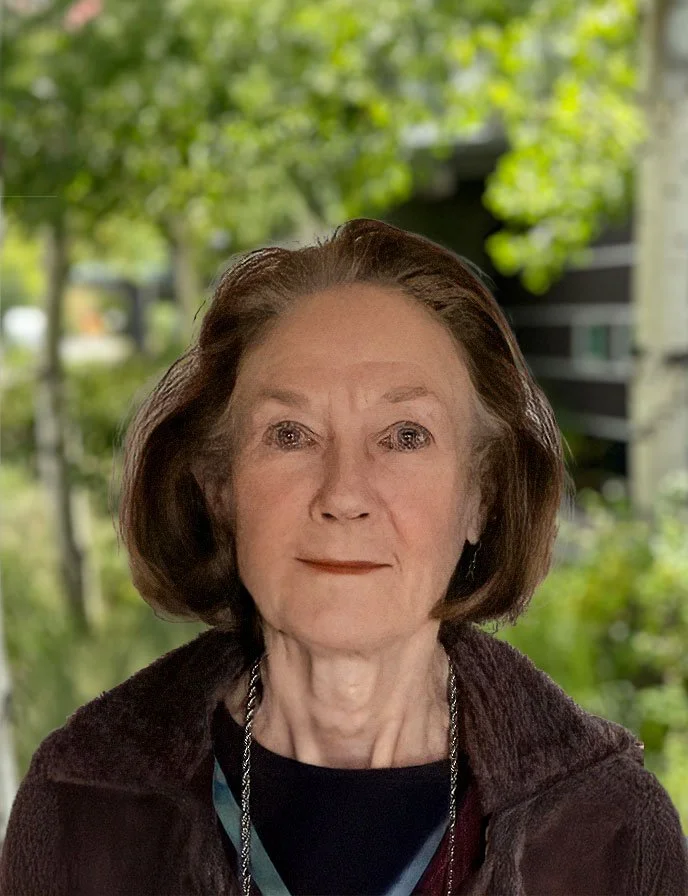 Close-up of an elderly woman with shoulder-length brown hair, wearing a black top and a brown jacket, outdoors with green trees in the background.