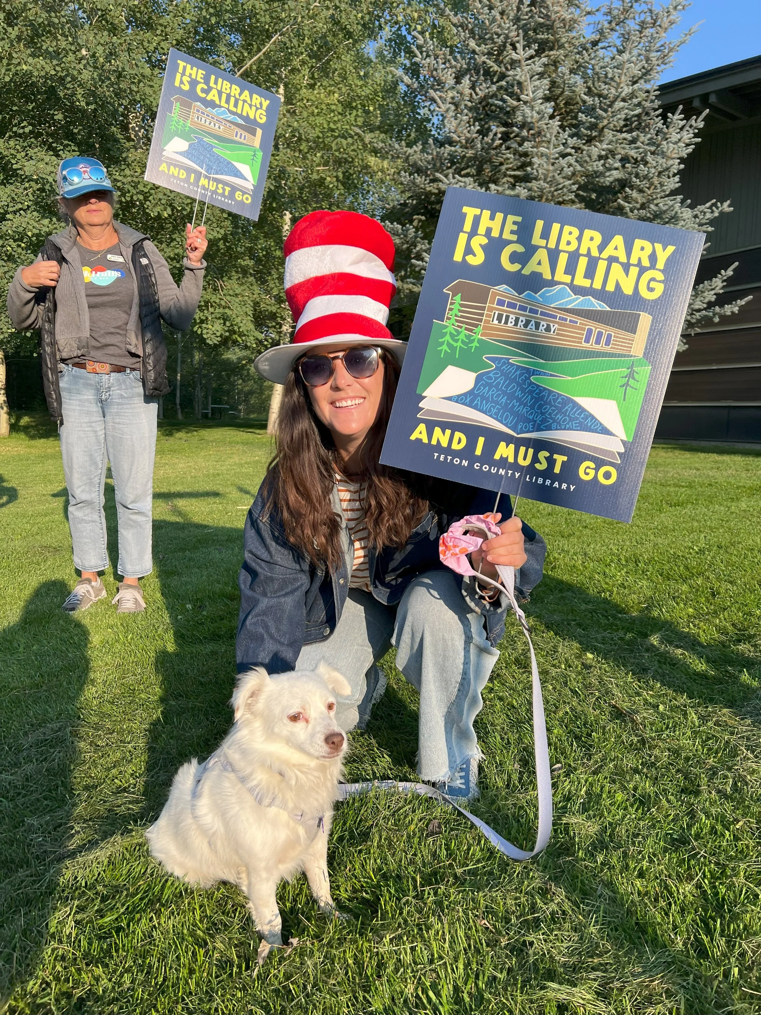 Two women holding signs that say 'The library is calling and I must go' standing on grass in a park with trees and a building in the background. One woman is kneeling with a small white dog, wearing sunglasses and a red and white striped hat resembli