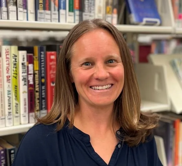 A woman with long light brown hair smiling at the camera in a library, with bookshelves filled with books in the background.