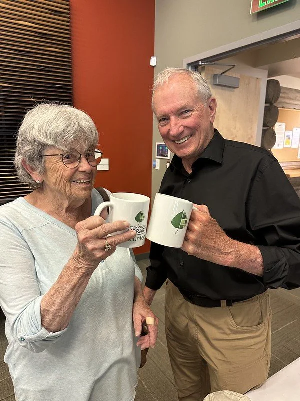 An elderly woman and an elderly man are smiling and holding white coffee mugs with a green leaf logo, standing indoors near a door.