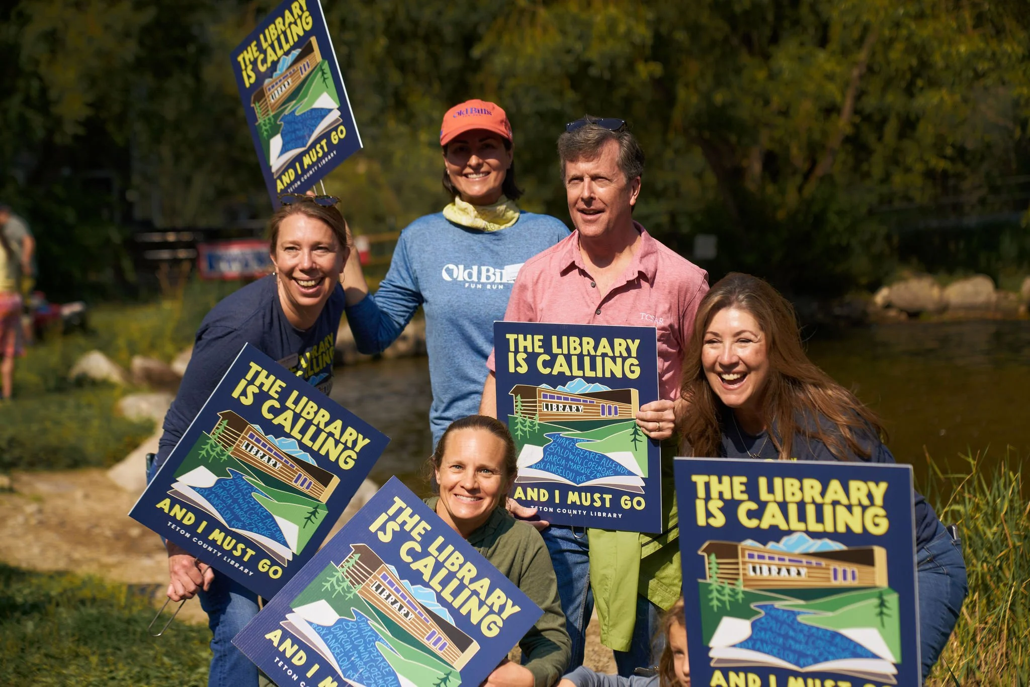 Group of people outdoors holding signs that read 'The library is calling and I must go.' The signs feature an illustration of a library building, a river, and mountains, with a colorful natural background.
