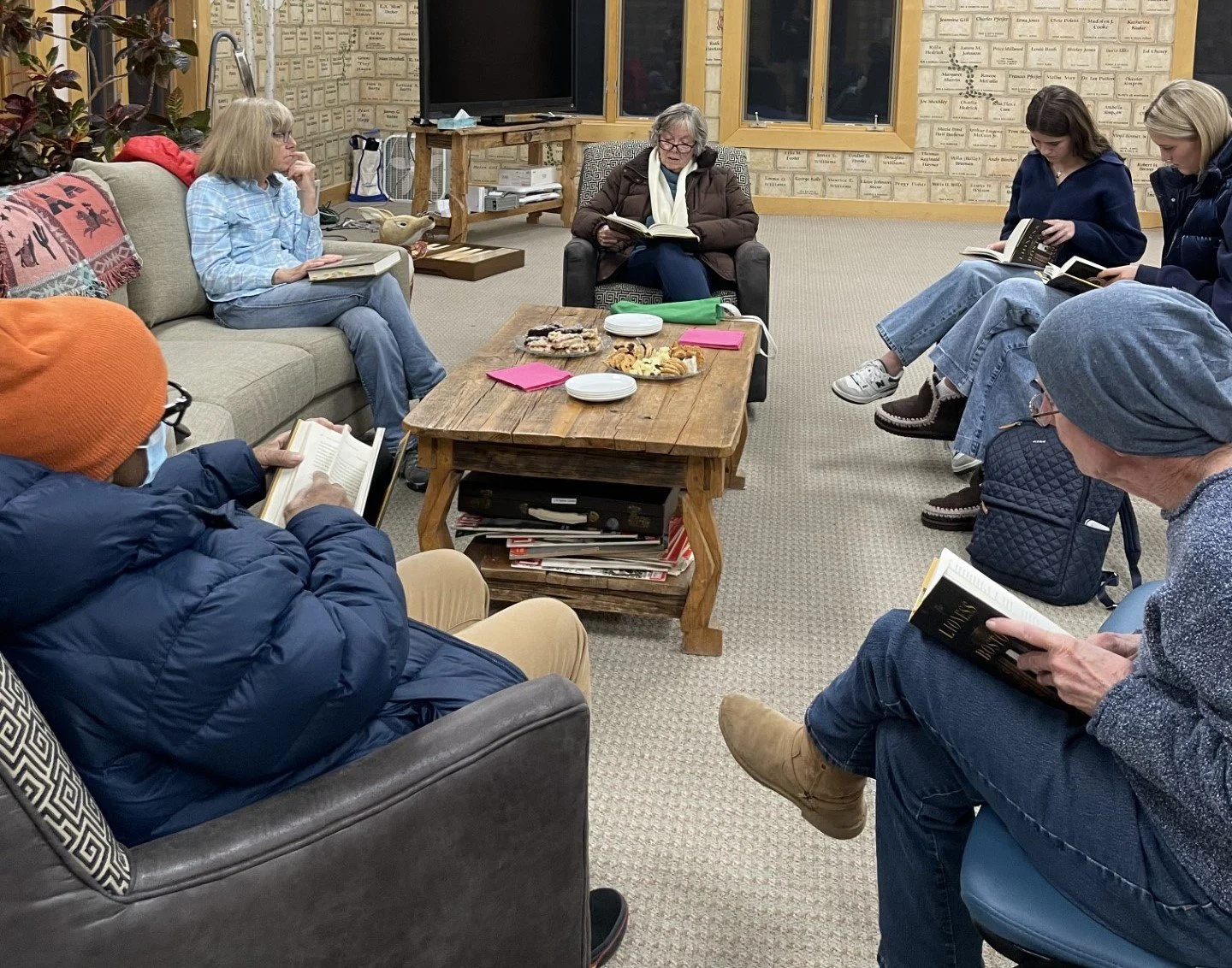 A group of people sitting in a circle in a cozy room, reading books, with a table of snacks in the center.