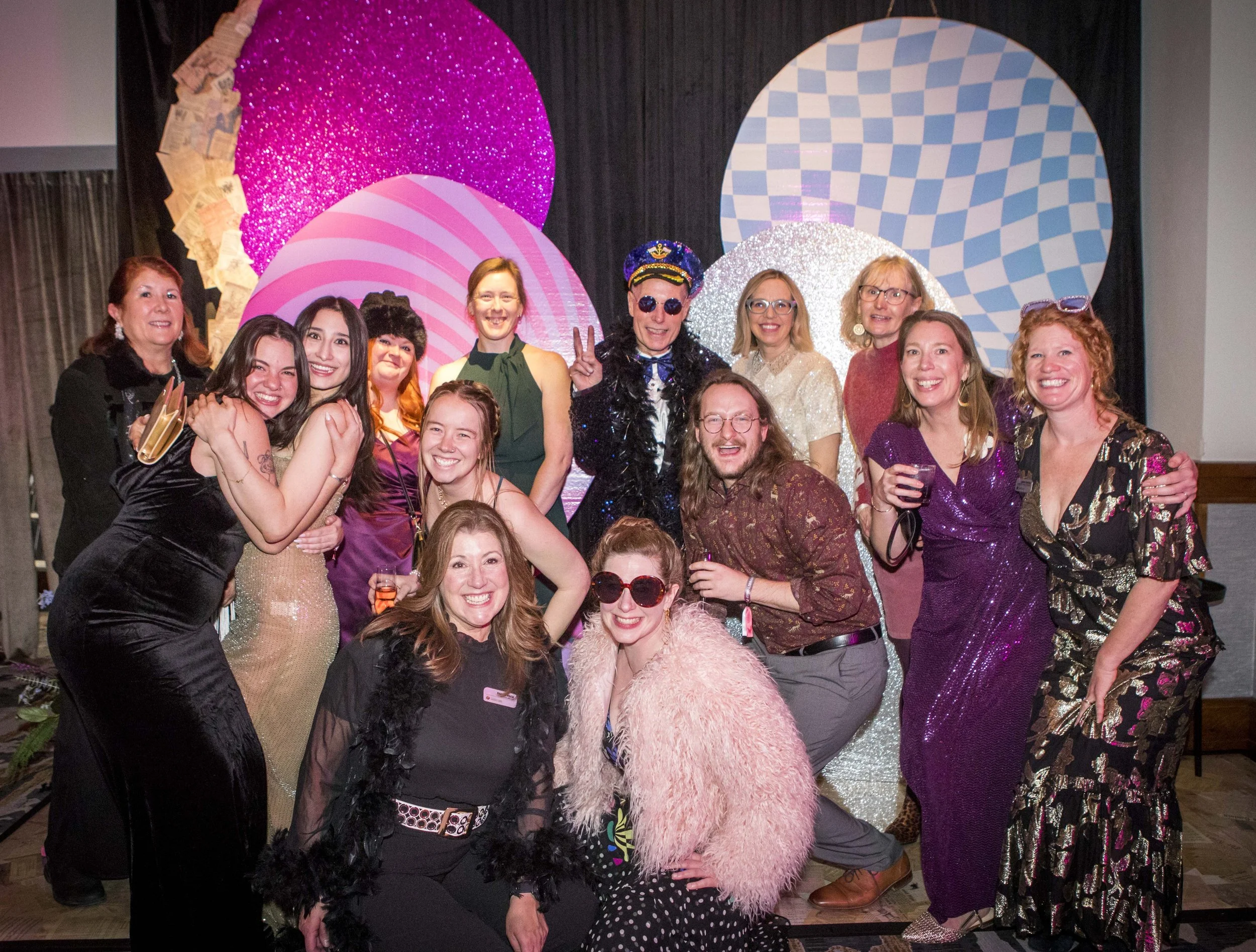 A group of people dressed in colorful and glamorous outfits, celebrating at a party with a vibrant, decorated backdrop featuring patterned circles and glitter.