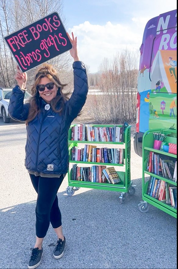 Woman smiling and holding a sign that says 'FREE BOOKS libras gratis' next to bookshelves with books outside.