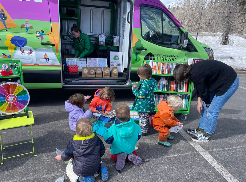 Children and adults gathered around a mobile library parked outdoors, with books and a colorful spinning wheel game visible.